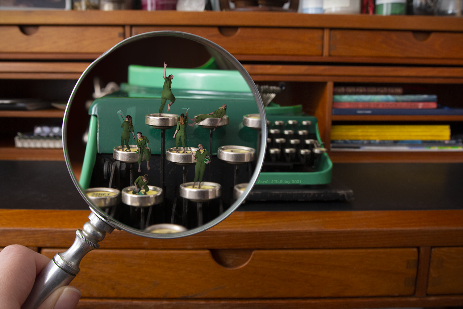 A hand holding a magnifying glass that enlarges miniature figures of people dressed in green scrubs, placed on the keys of a vintage typewriter, with a wooden desk and shelves with books and jars in the background.