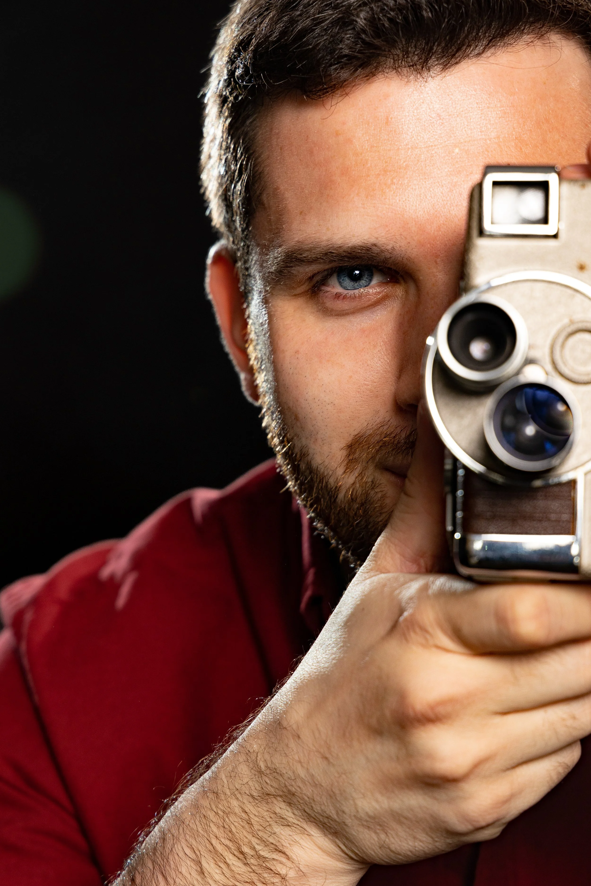 A man with short dark hair and a beard taking a close-up selfie with an old-fashioned film camera, wearing a red shirt, against a dark background.