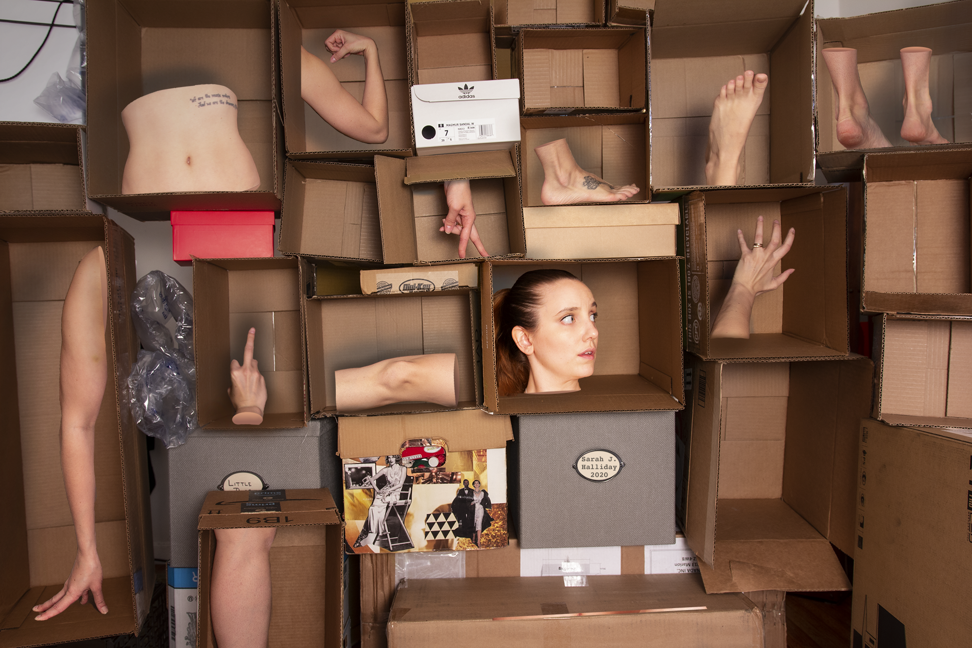 Art installation featuring cutouts of human body parts, including arms, legs, a torso, and a head, arranged within cardboard boxes and shelves.