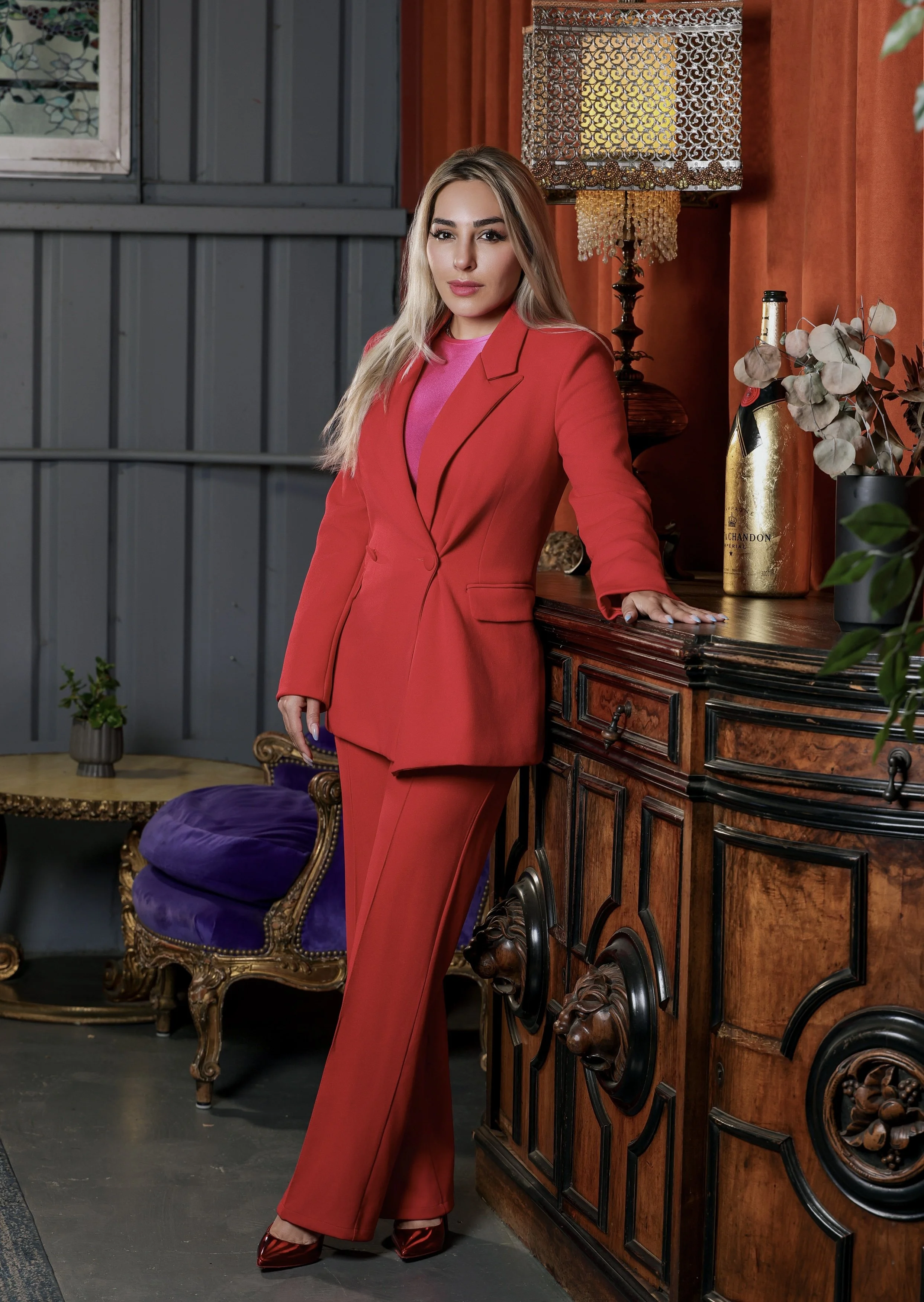 A woman with long blonde hair wearing a red blazer and matching red pants, standing indoors beside an ornate wooden sideboard, with her hand resting on the surface. The room has a vintage decor with an orange curtain, a decorative lamp with a beaded fringe, and bottles of champagne. There is also a purple velvet armchair with gold trim and a small potted plant in the background.