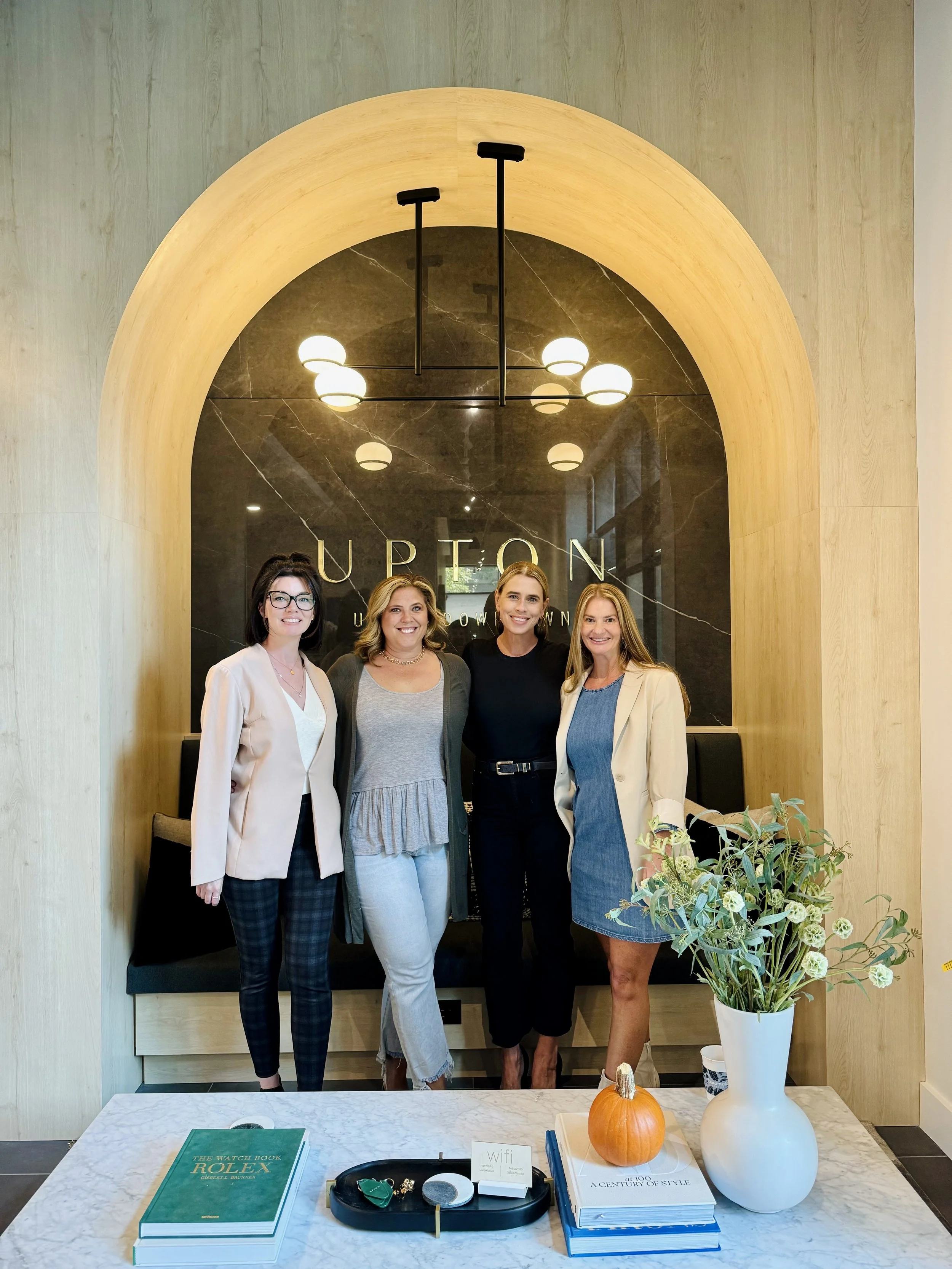 Four women standing together inside a stylish, modern interior with a black marble wall, large arched wooden frame, and warm lighting, in front of a sign that reads 'UPTON'. A marble table in the foreground holds a green book, a small pumpkin, a whit