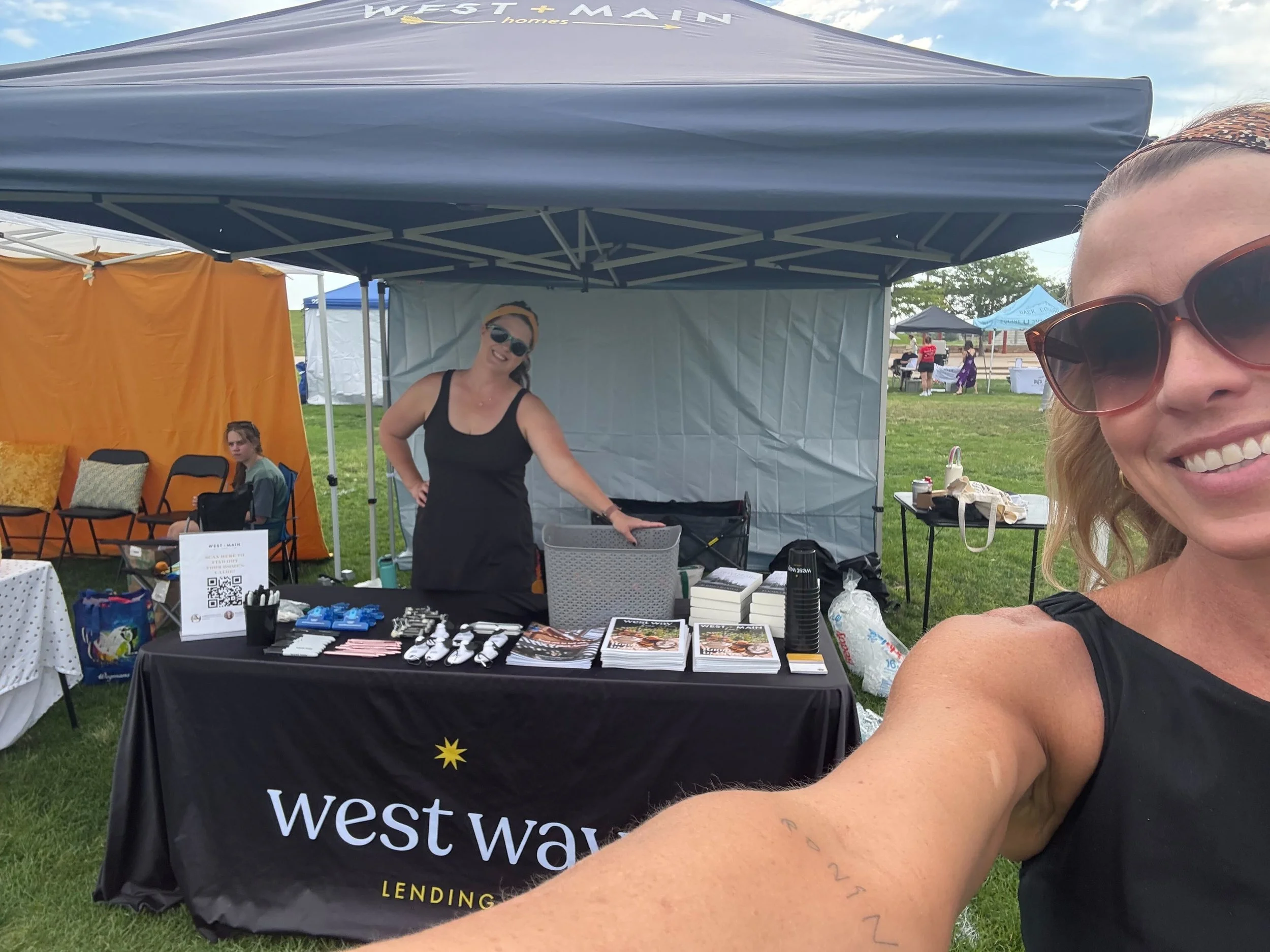 Smiling woman with sunglasses taking a selfie in front of a booth labeled 'Westway Lending,' with another woman standing behind the booth, under a canopy at an outdoor event.