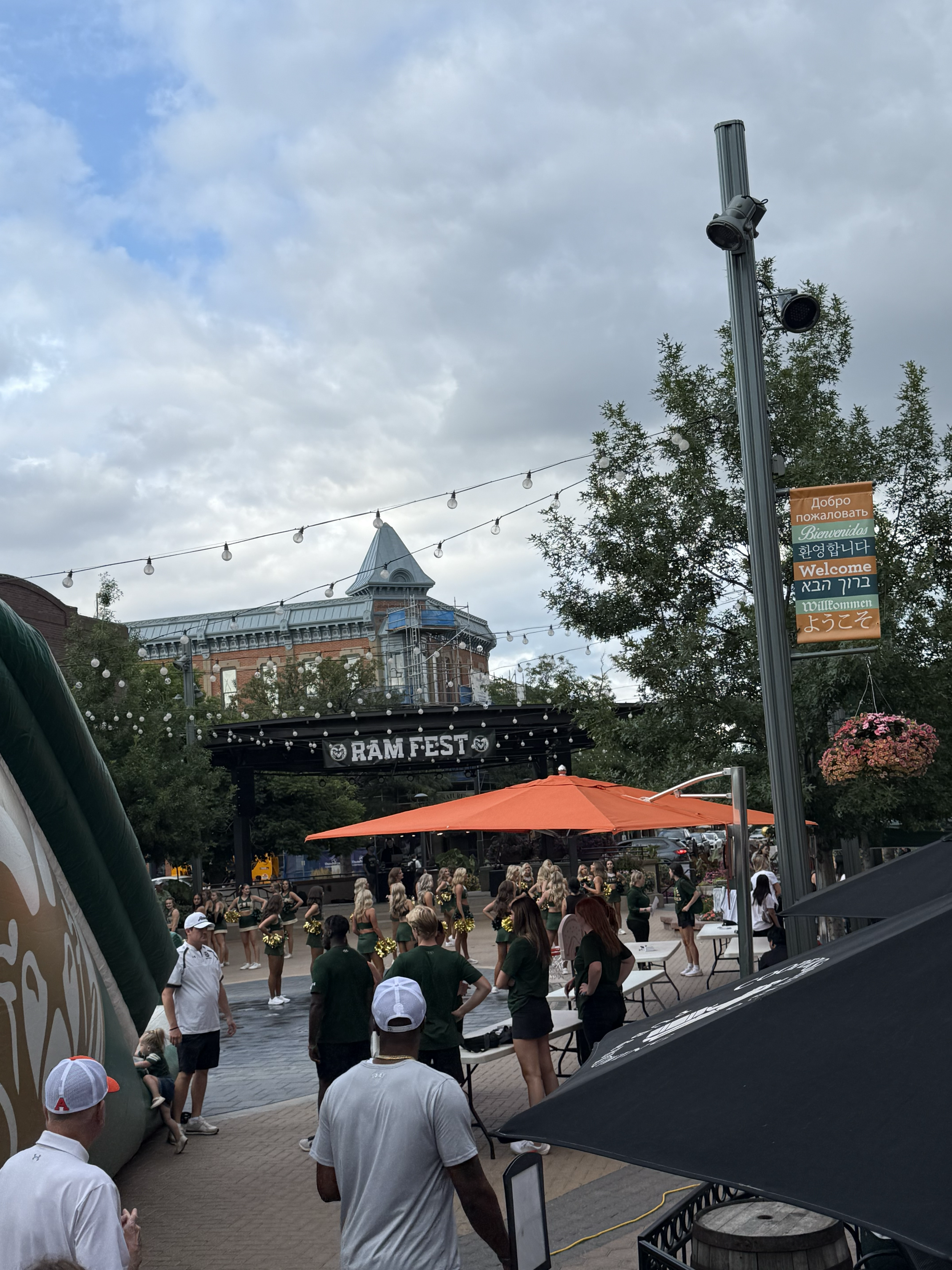 People gathered at RAM Fest with a stage, tents, and string lights in an outdoor urban setting during daytime, with a historic building and trees in the background.