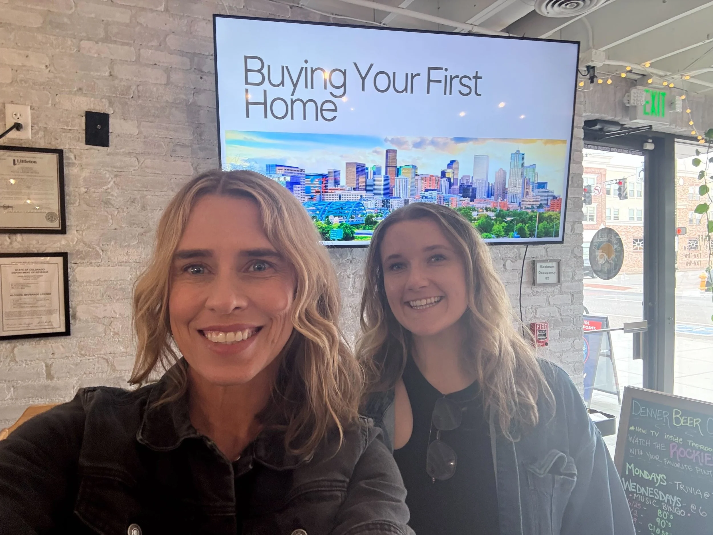 Two women smiling in a restaurant with a view of the city skyline on a TV screen behind them. The TV screen displays the text 'Buying Your First Home'. The restaurant has a brick wall interior and large windows.