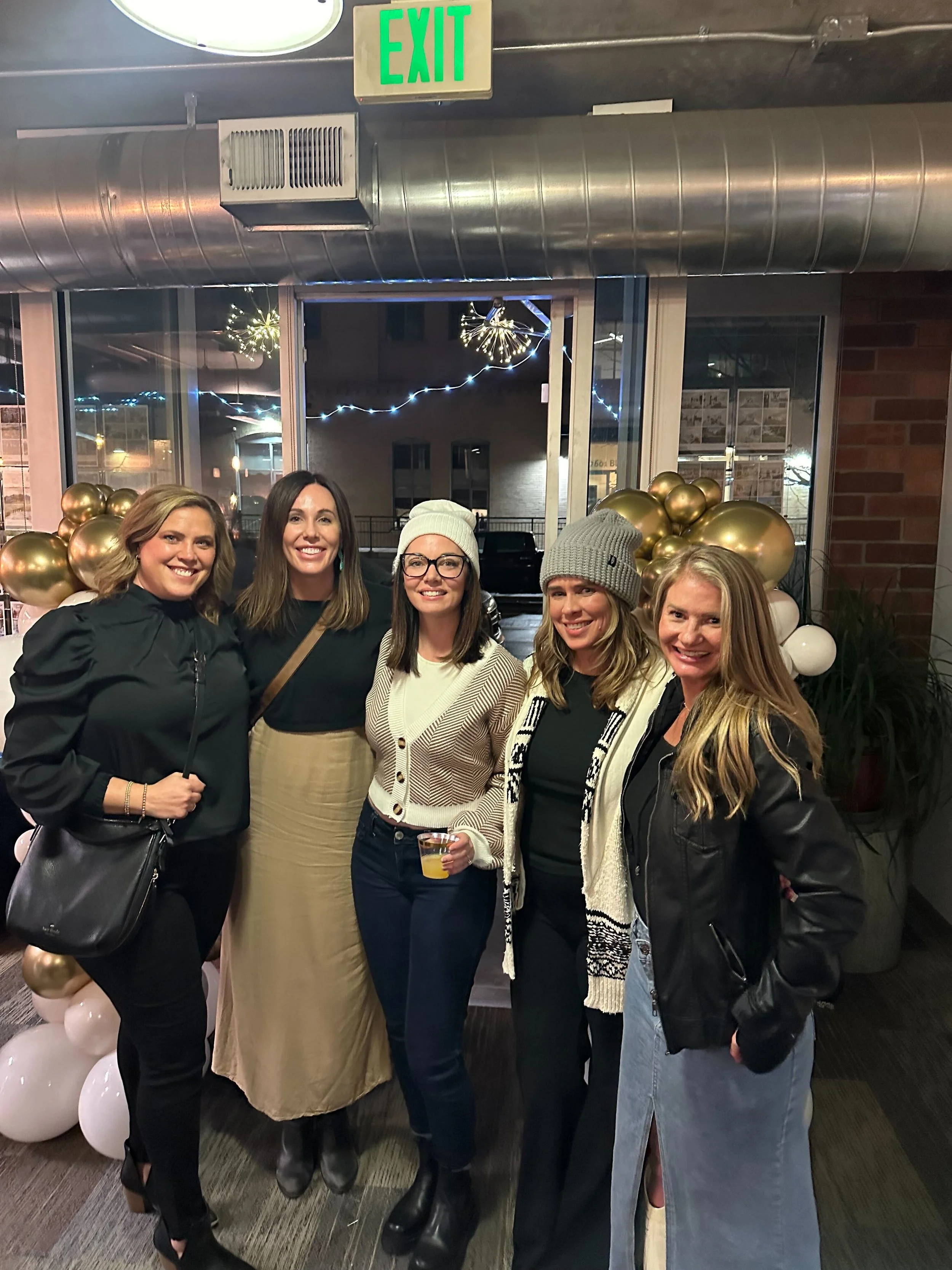 Five women standing together at a social event, smiling, in front of a window decorated with balloons and string lights.