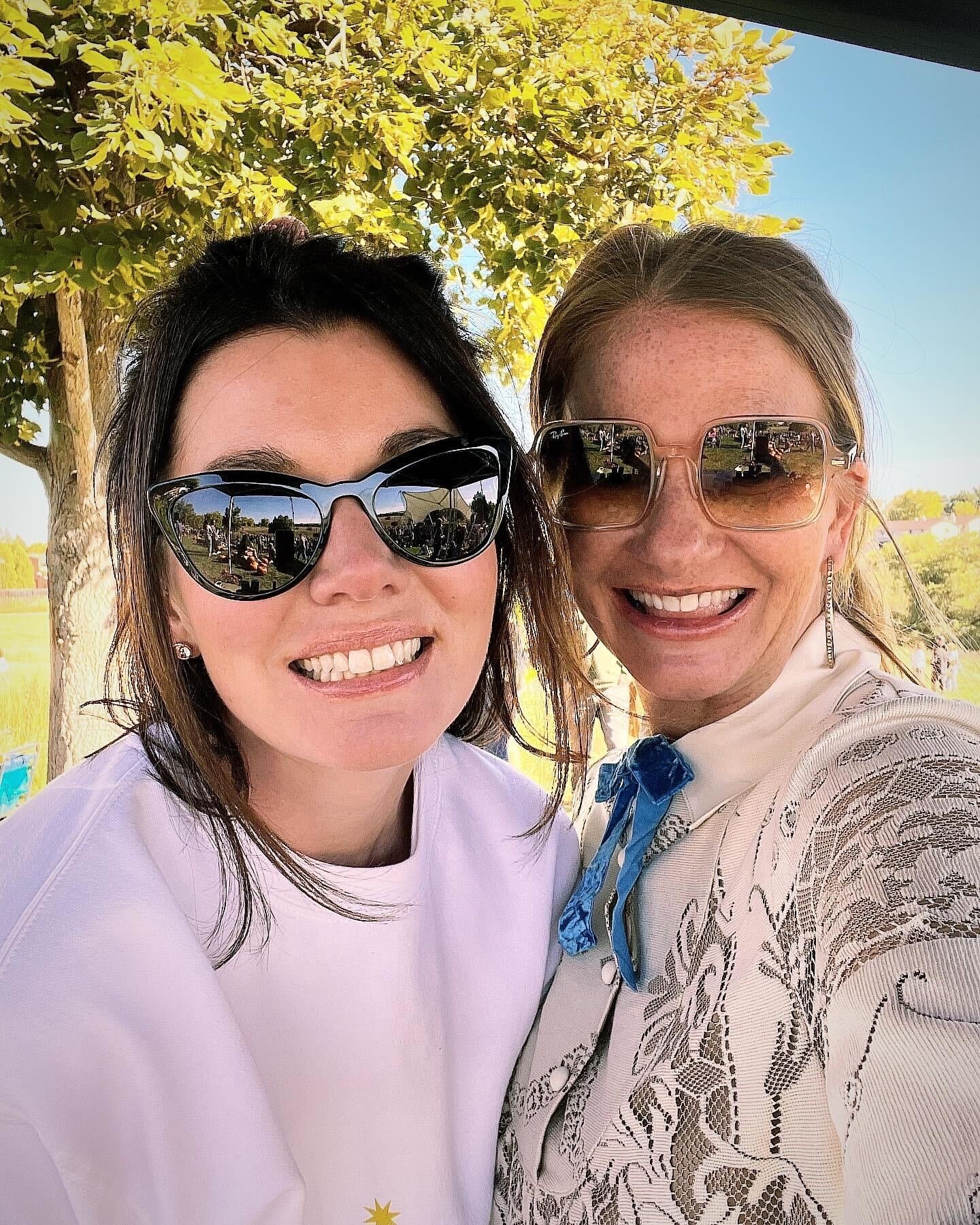 Two women wearing sunglasses smiling outdoors with a tree and bright blue sky in the background.