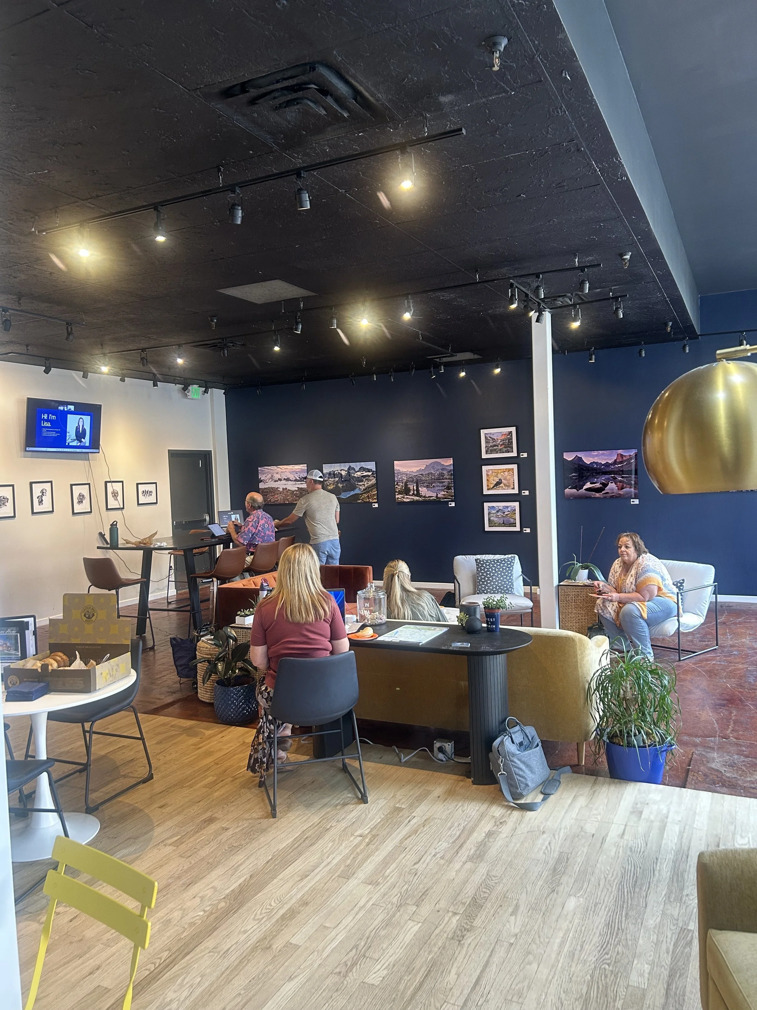 Art gallery or cafe with people sitting and socializing, dark blue accent walls with framed landscape photographs, modern seating, a large gold hanging lamp, and a television screen displaying a presentation or slideshow.