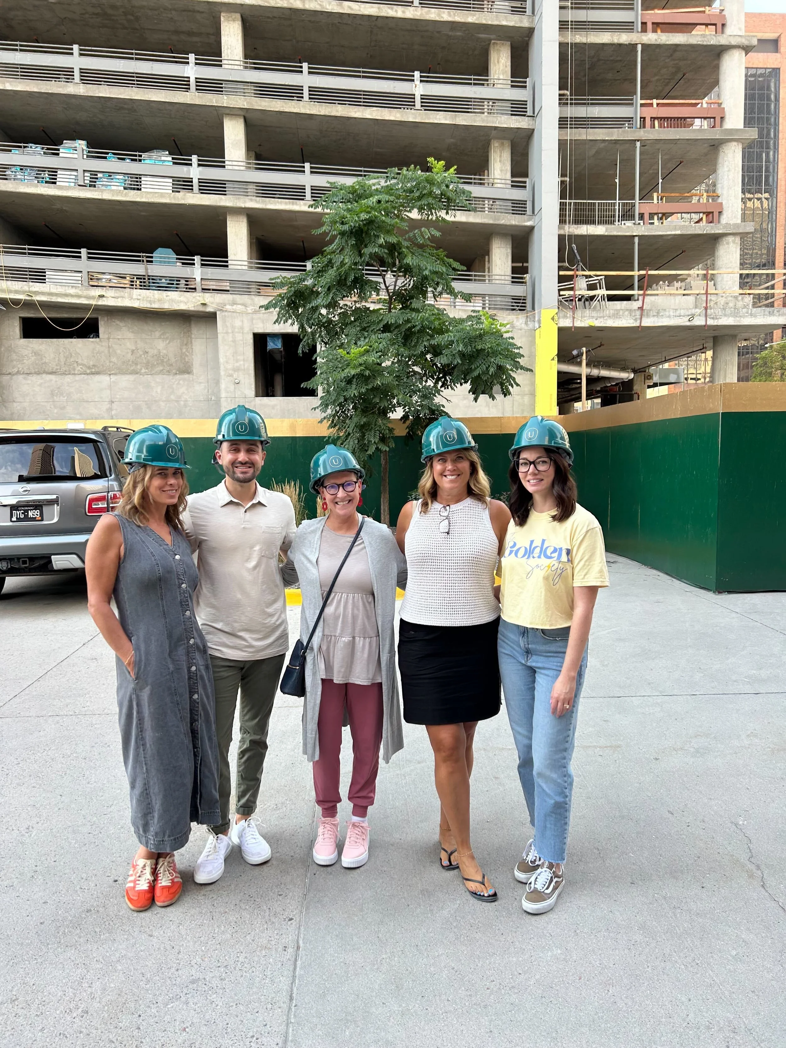 Five women standing outdoors in front of a construction site with a partially built multi-story building, wearing blue safety helmets.