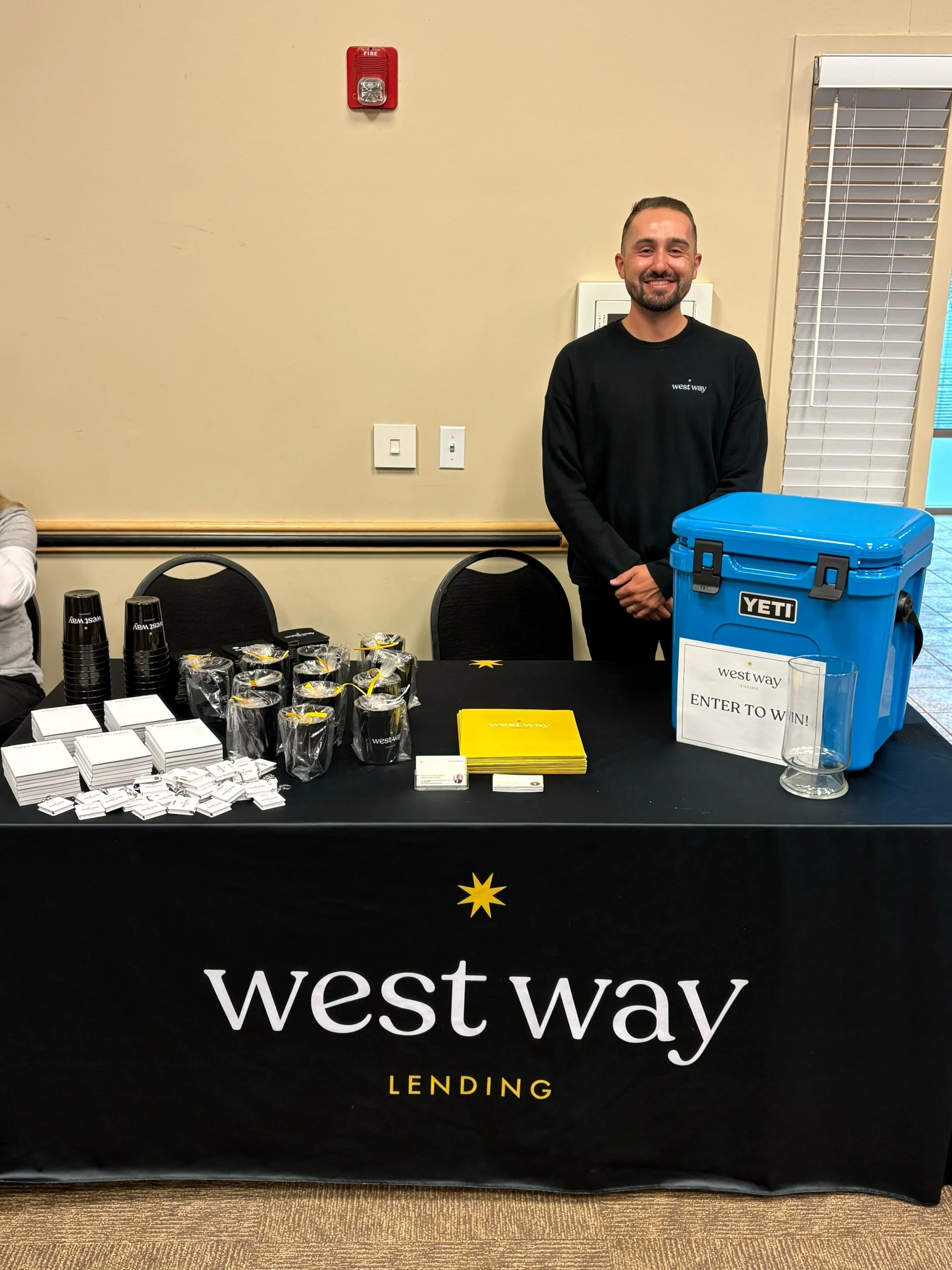 A man standing behind a table with branding that reads 'West Way Lending'. The table has promotional items including a yellow brochure, white notepads, pens, and a blue YETI cooler with a sign that says 'Enter to Win'. There are black cups and small 