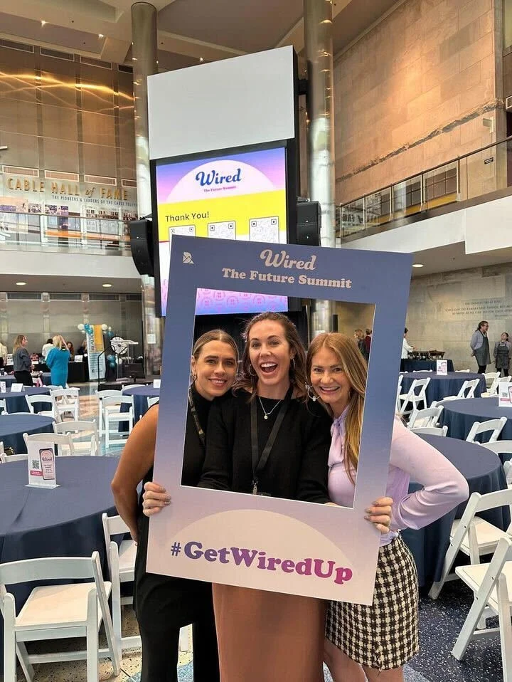 Three women smiling and posing inside a large rectangular frame with the hashtag #GetWiredUp at a Wired event called The Future Summit, with tables and chairs in a large, modern indoor venue.