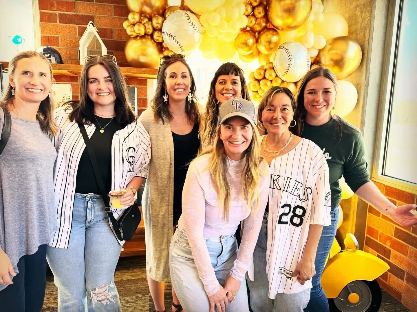 Group of eight women smiling at baseball-themed celebration with yellow, gold, and white balloons, including baseball and baseball glove decorations.