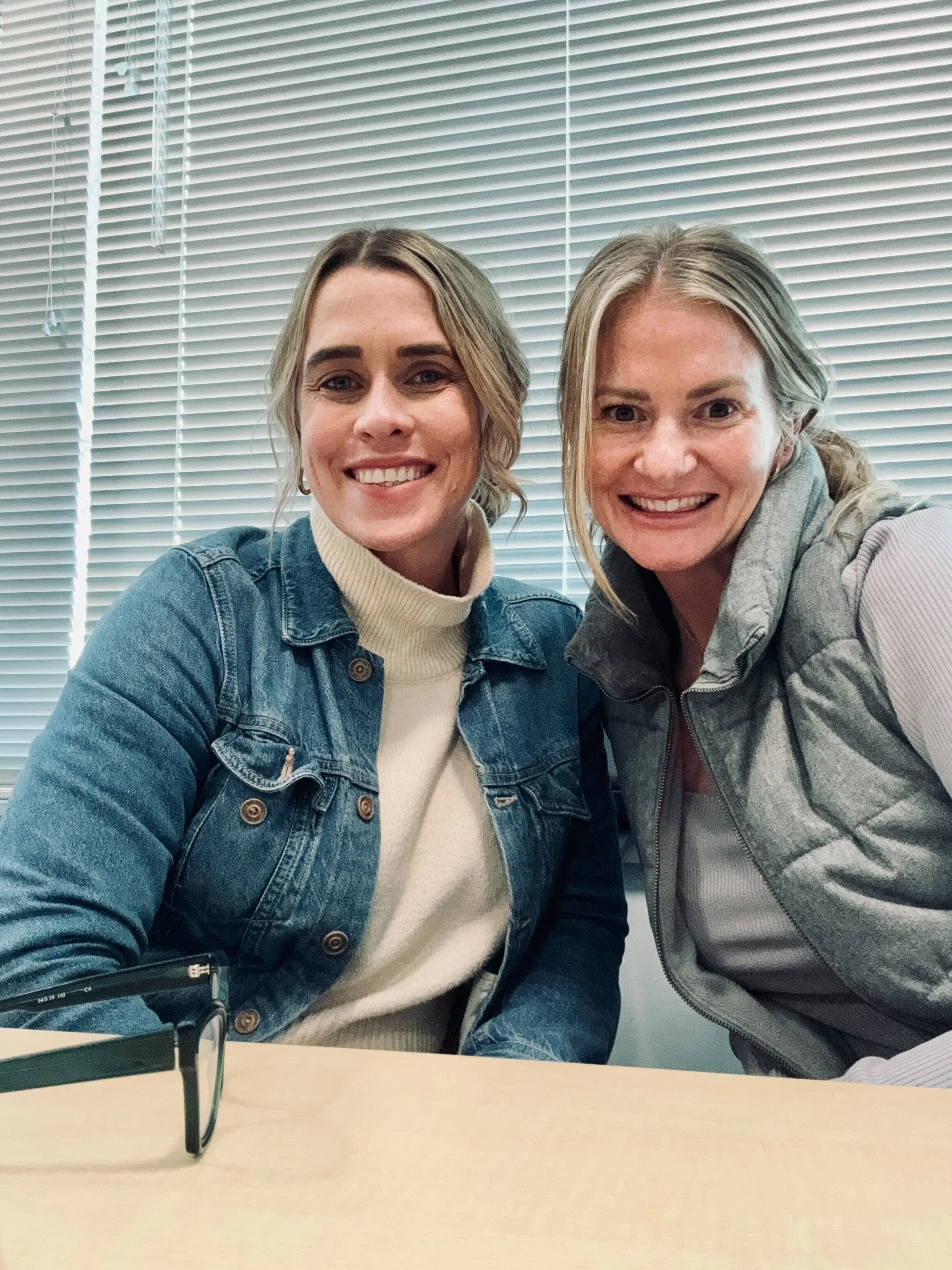 Two women smiling, sitting in front of horizontal window blinds in an office or meeting room.