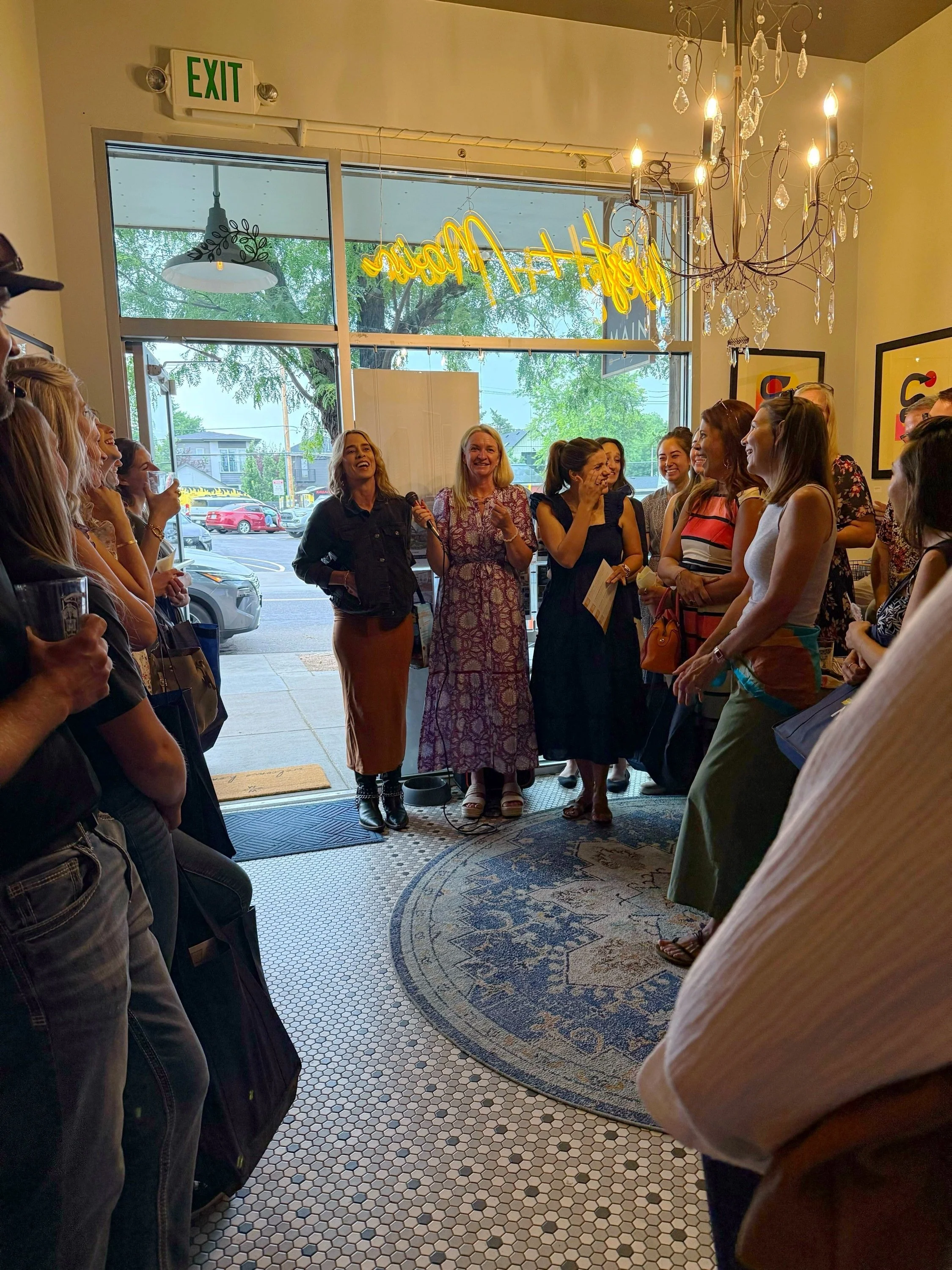 A group of women gathered inside a cafe, listening to a woman speaking with a microphone. There is a window with a view outside, a chandelier hanging from the ceiling, and a neon sign that reads 'hello my' in yellow. The atmosphere appears lively and
