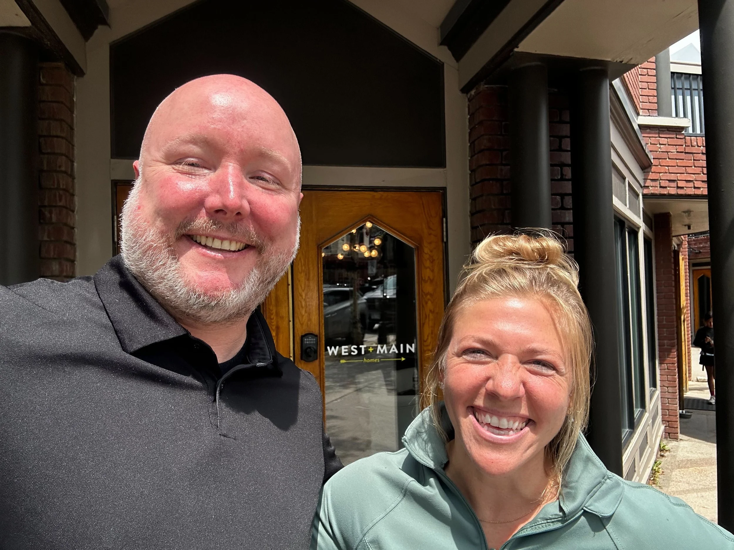 Two smiling people, a man with a beard and a woman, are taking a selfie outside a brick building with a wooden door marked 'WEST + MAIN'.