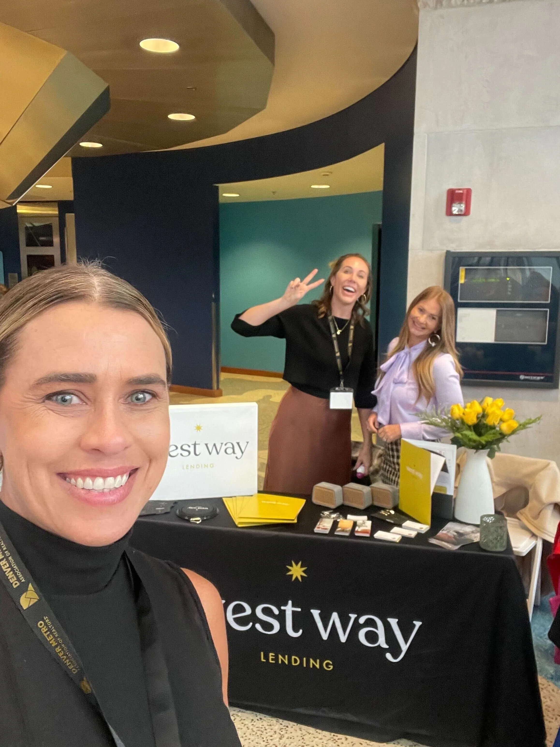 A woman taking a selfie at a Westway Lending booth with two other women smiling behind the table, which has a sign, flowers, and promotional materials.