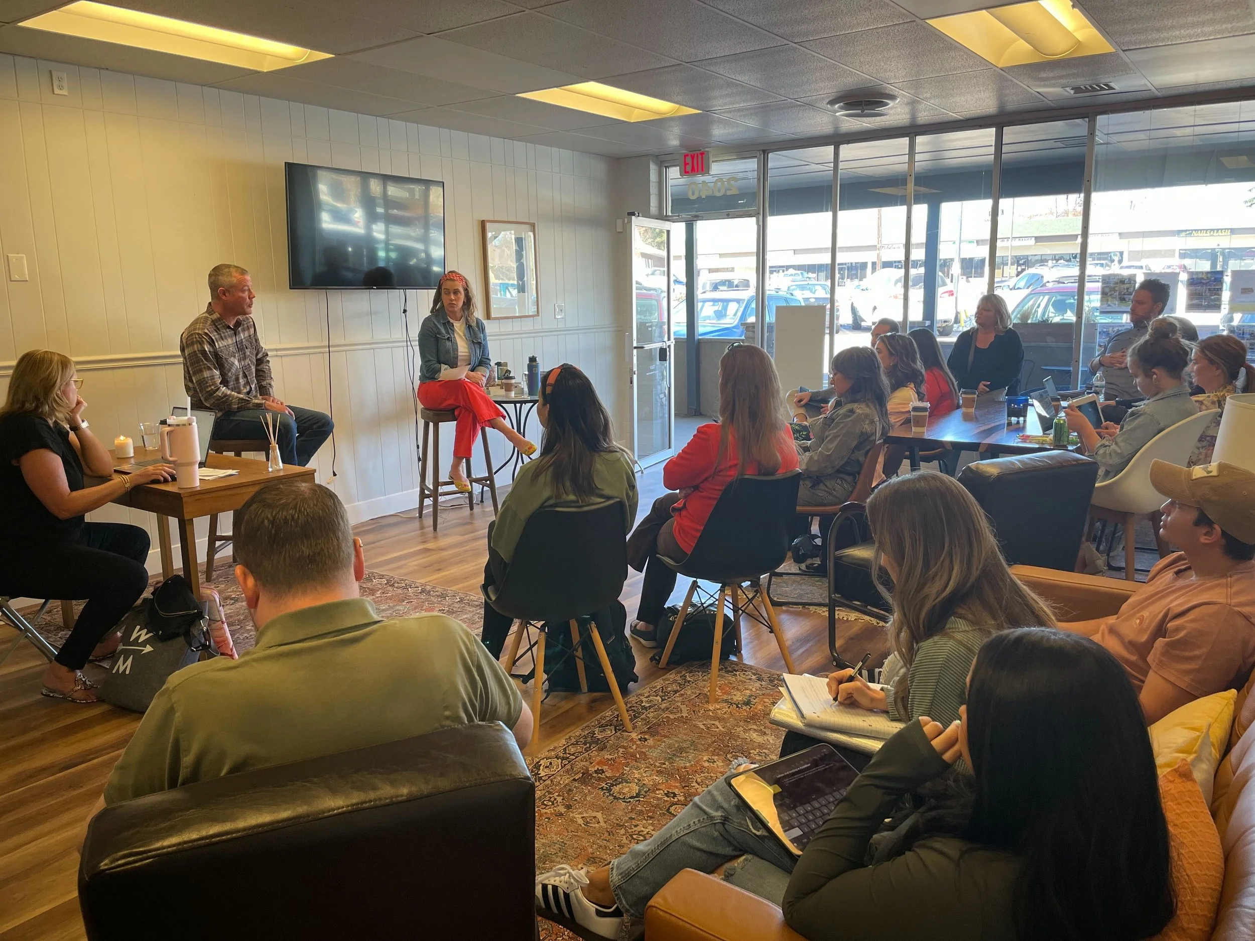 A group of people attending a presentation or discussion in a cozy, well-lit room with large windows showing parked cars outside. Two speakers are seated on stools at the front near a large television screen, engaging with the audience.