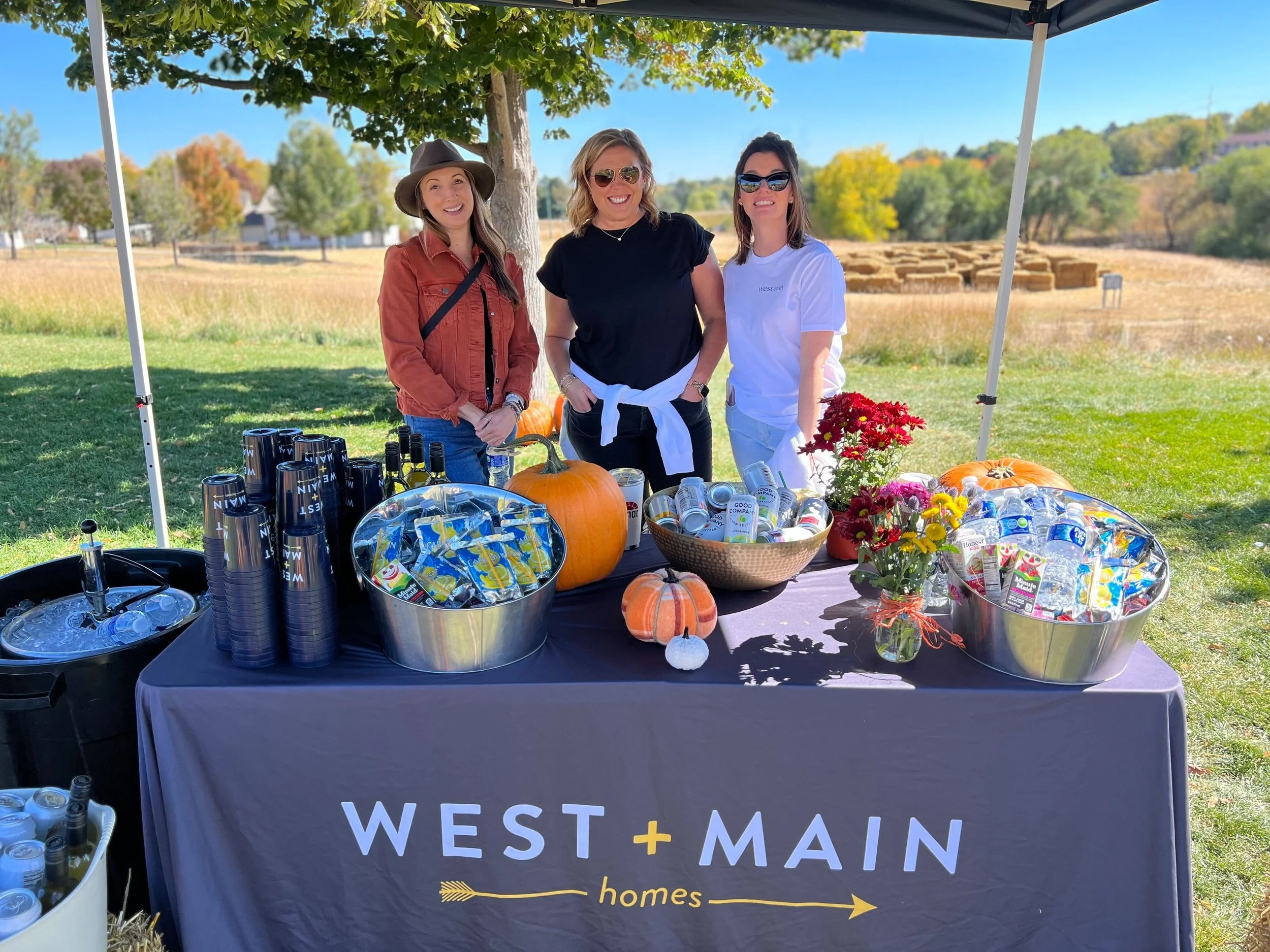 Three women standing behind a table under a canopy at an outdoor event on a sunny day. The table has a West + Main Homes tablecloth and is decorated with pumpkins, flowers, and beverages. In the background, there are trees and a field.