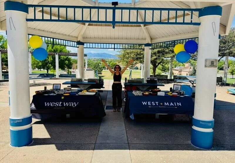 A smiling woman wearing sunglasses standing under a pavilion with arms raised. Two tables covered with black tablecloths labeled 'west way' and 'WEST + MAIN' are set up beside her, with balloons and promotional materials. Park and trees are visible i