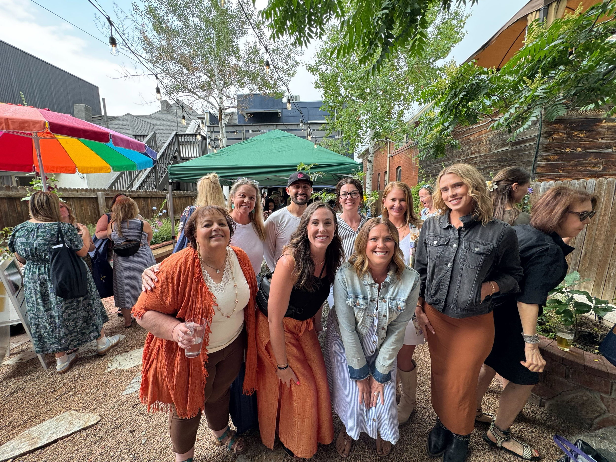 Group of smiling women and a man at an outdoor gathering, some holding drinks, in a backyard with colorful umbrellas and string lights.