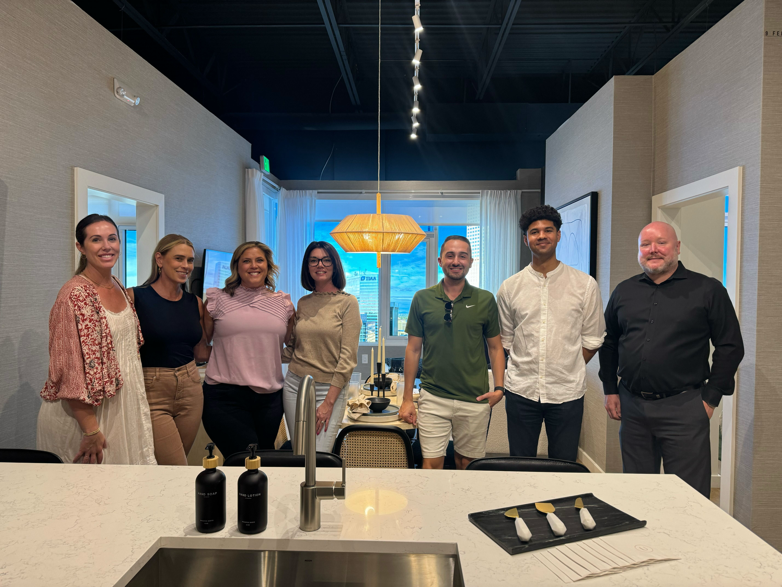 Seven people standing in a modern, well-lit kitchen with a view of a cityscape outside the window.