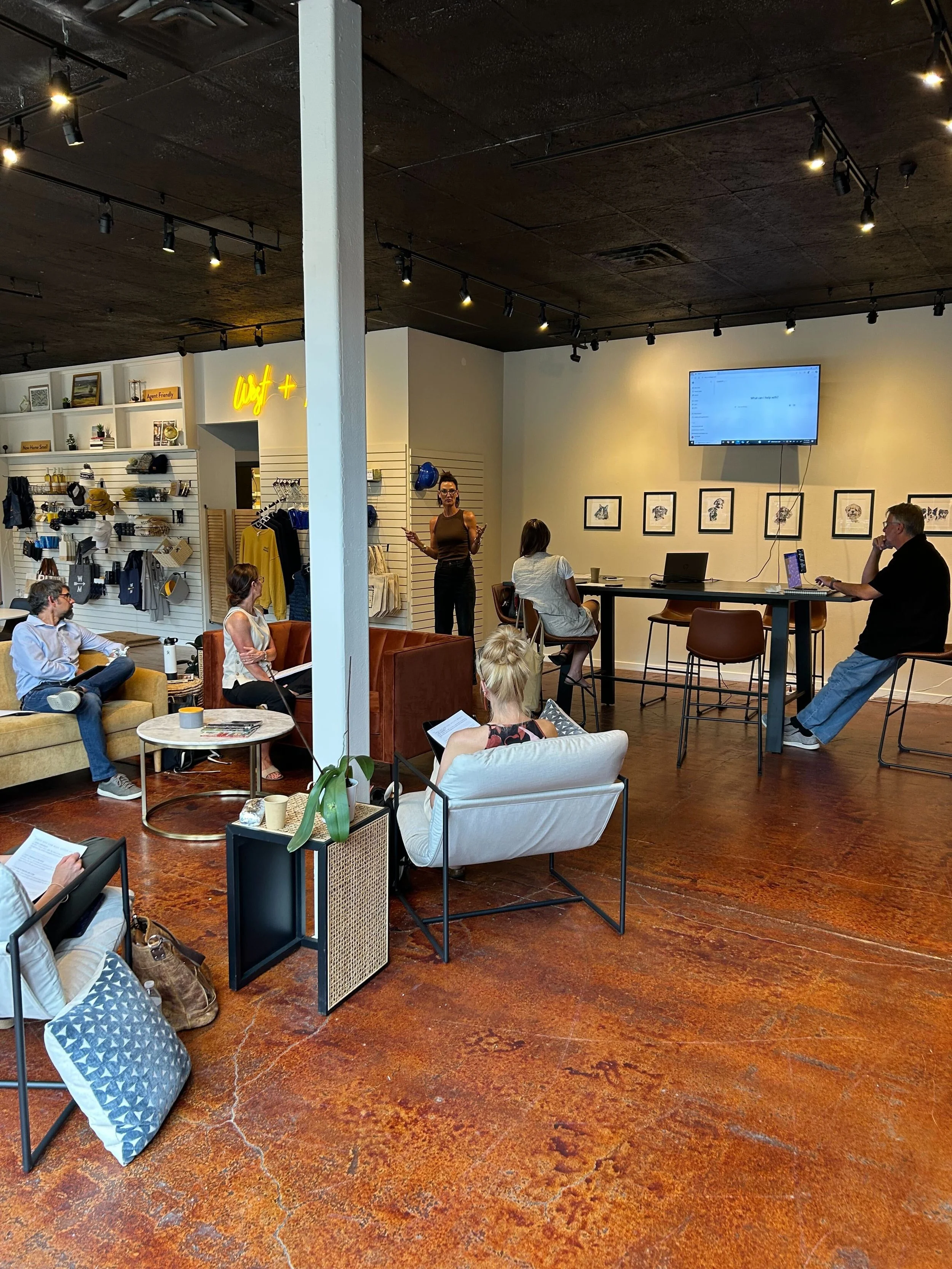 People attending a presentation or workshop in a modern, industrial-style space with exposed ceiling, wooden floor, and shelves holding various items. A woman stands near the wall speaking, while others sit on chairs and sofas, some using laptops or 