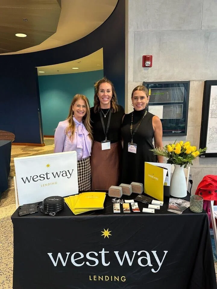 Three women standing behind a table with 'West Way Lending' branding at an indoor event. The table has brochures, small speakers, a vase with yellow flowers, and other promotional materials.