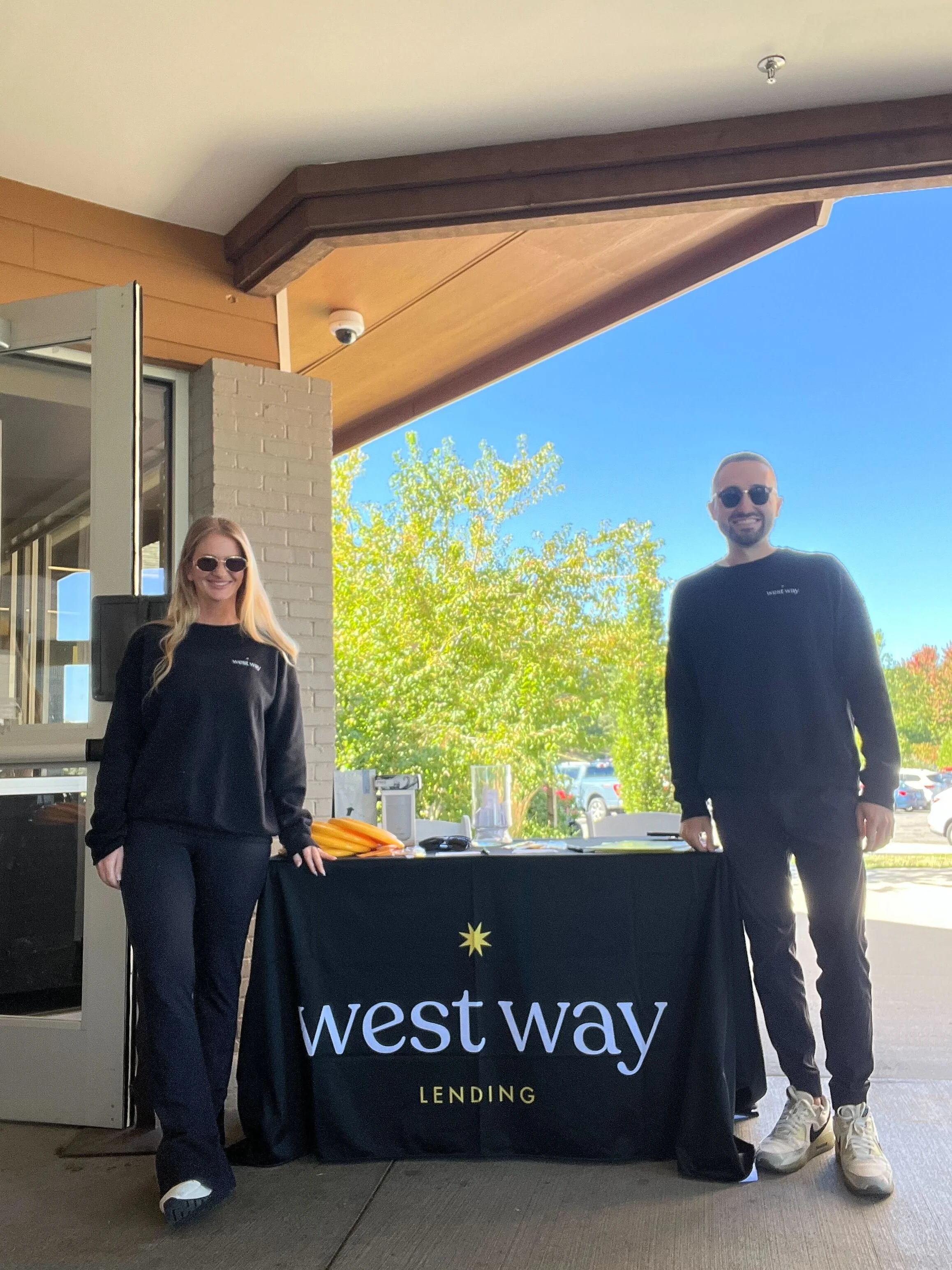 Two people standing behind a table with a 'West Way Lending' banner outdoors on a sunny day. The woman on the left has long blonde hair, is wearing sunglasses, a black sweatshirt, and black pants. The man on the right has short hair, is wearing sungl