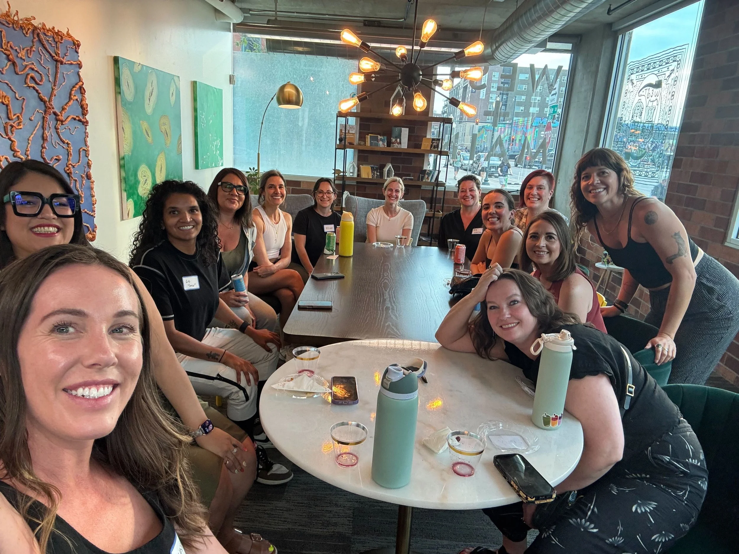 Group of women sitting and standing around a large shared table in a modern, art-filled cafe or restaurant, smiling at the camera.