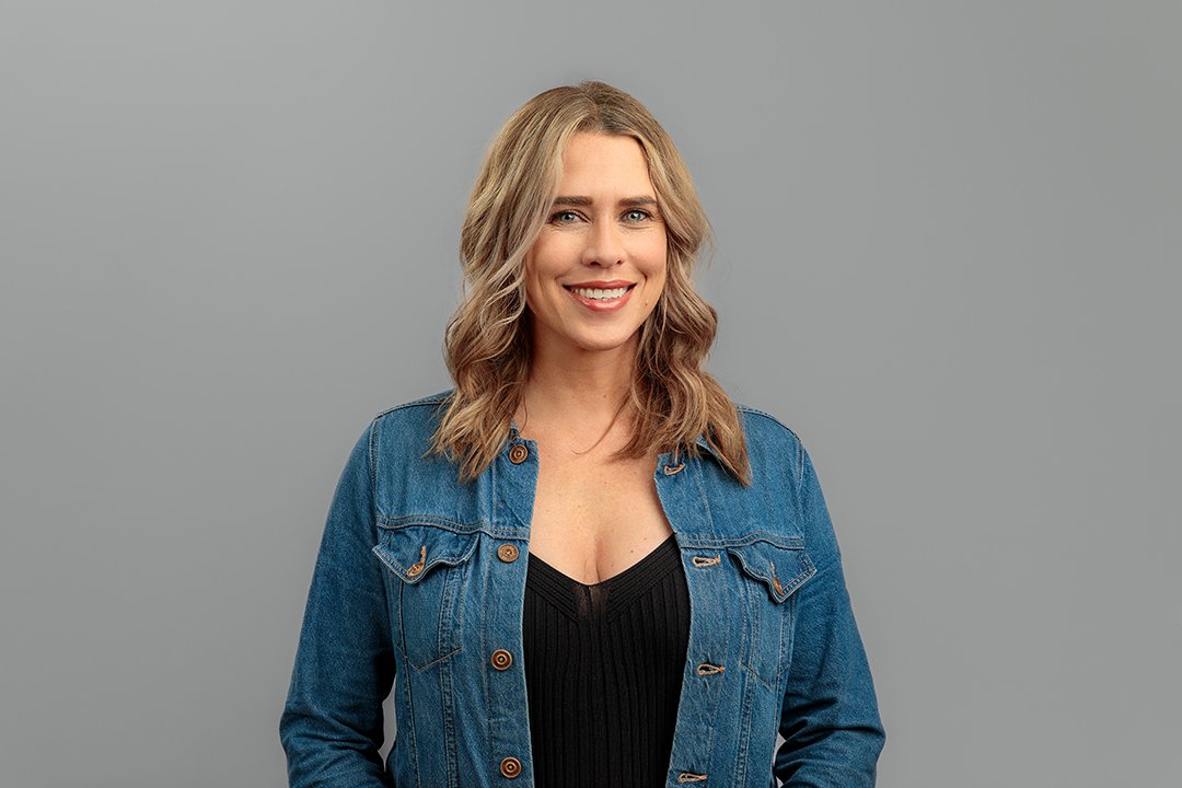 A woman with blonde hair, smiling, wearing a denim jacket over a black top, standing against a plain gray background.