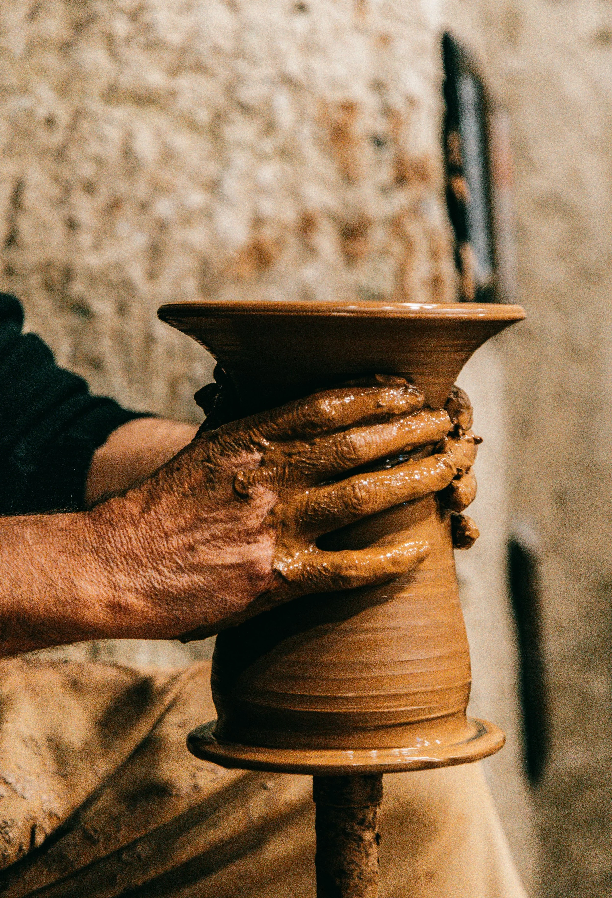 A person’s hand covered in clay, holding a pottery wheel with a spinning clay vessel. The background shows a textured stone wall.
