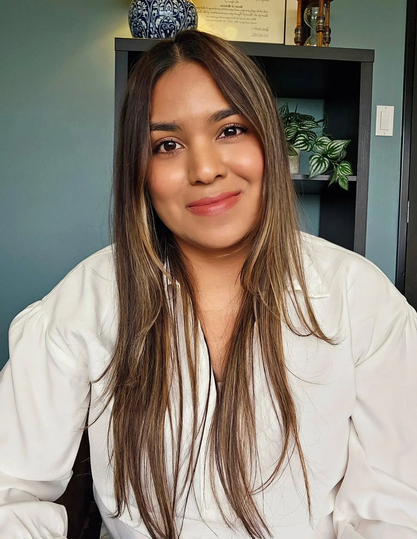 A woman with long, brown hair and a warm smile, wearing a white blouse, in a room with a dark bookshelf containing plants, a framed certificate, and decorative items, with teal-colored walls.
