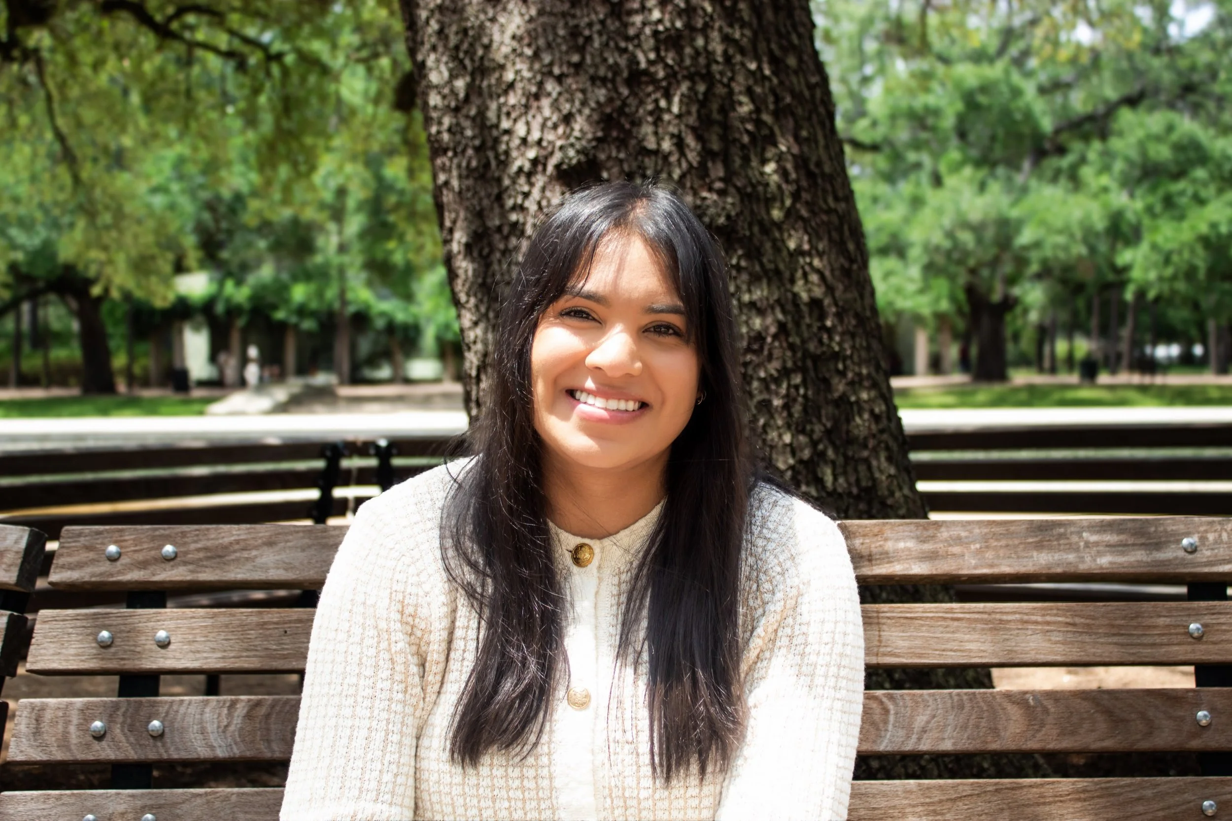 Smiling woman with long dark hair sitting on a park bench with trees and greenery in the background.