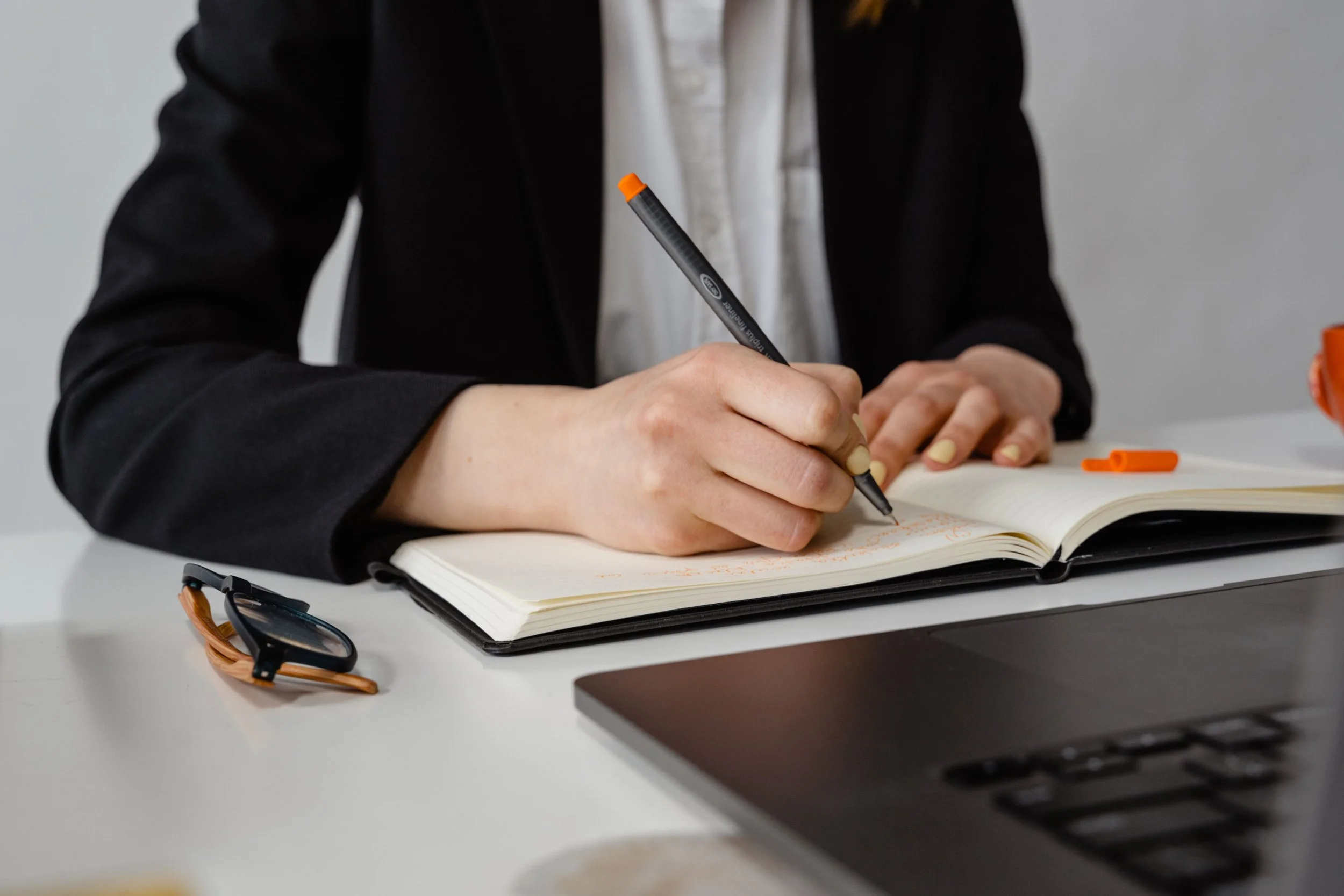 Person in a black blazer writing in a notebook with a black pen, on a white desk with sunglasses and a laptop.