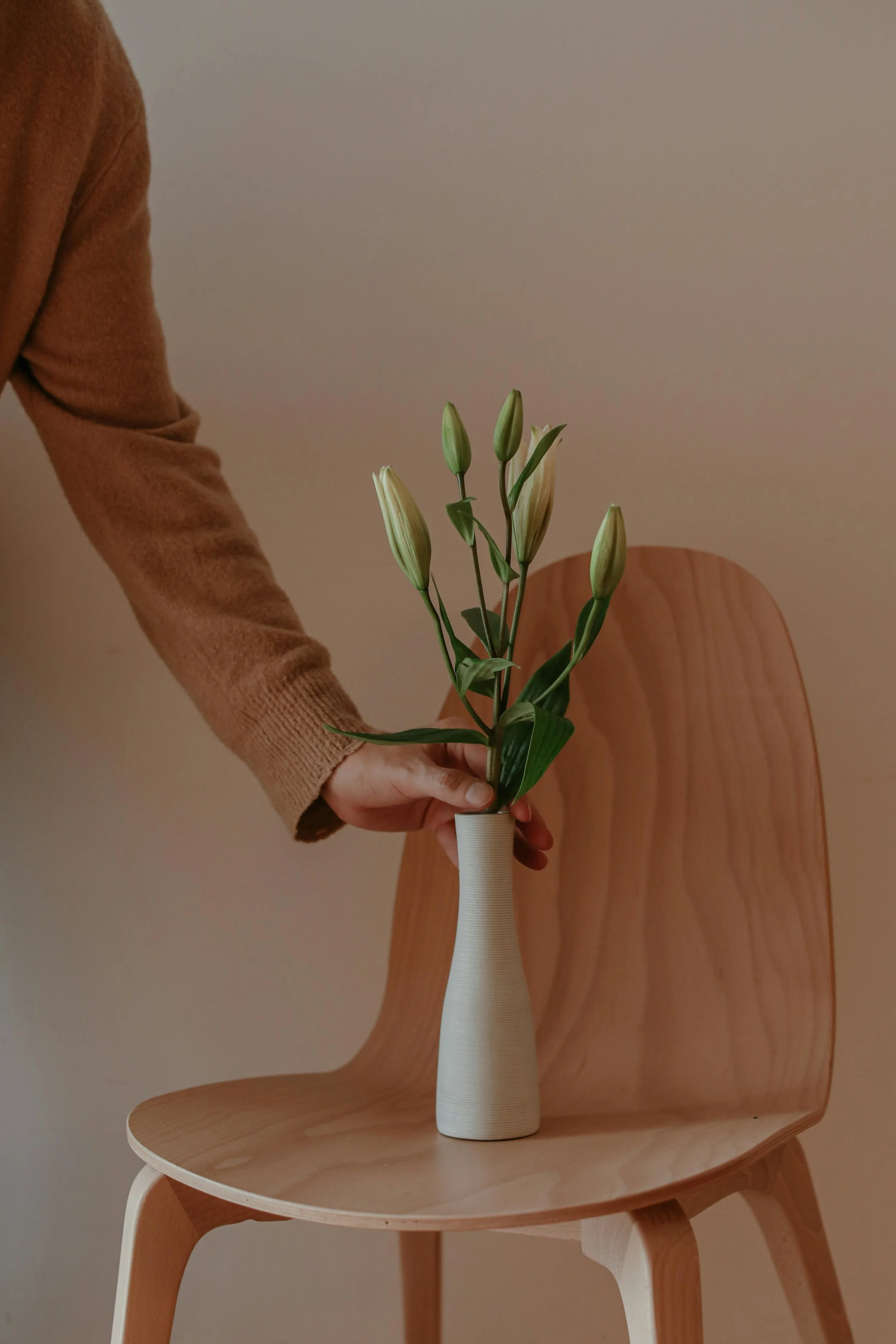 Person holding a white vase with white lilies, placed on a wooden chair with a light-colored wall in the background.
