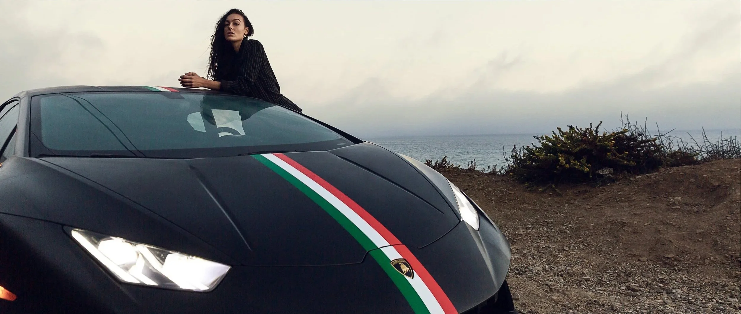 A woman with long dark hair leaning on the hood of a black Lamborghini sports car with an Italian flag stripe, near the ocean on a cloudy day.