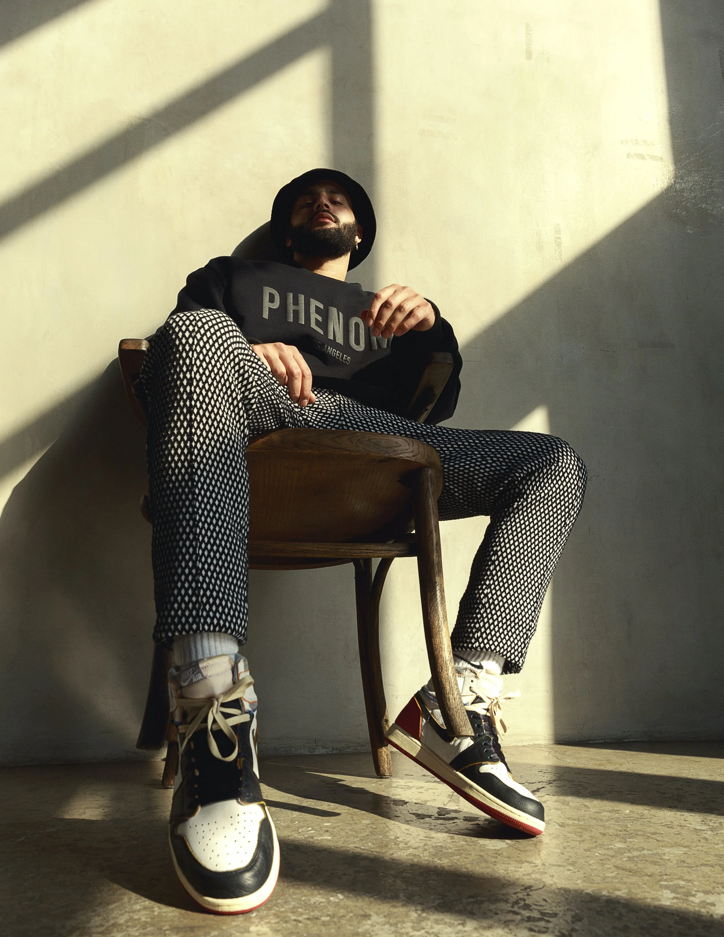 A person sitting on a wooden chair with legs spread apart, wearing black and white patterned pants, a black hoodie with text, white sneakers, and a black hat. The person's head is leaning back against the wall, and their face is partially illuminated by sunlight casting shadows on the wall behind them.