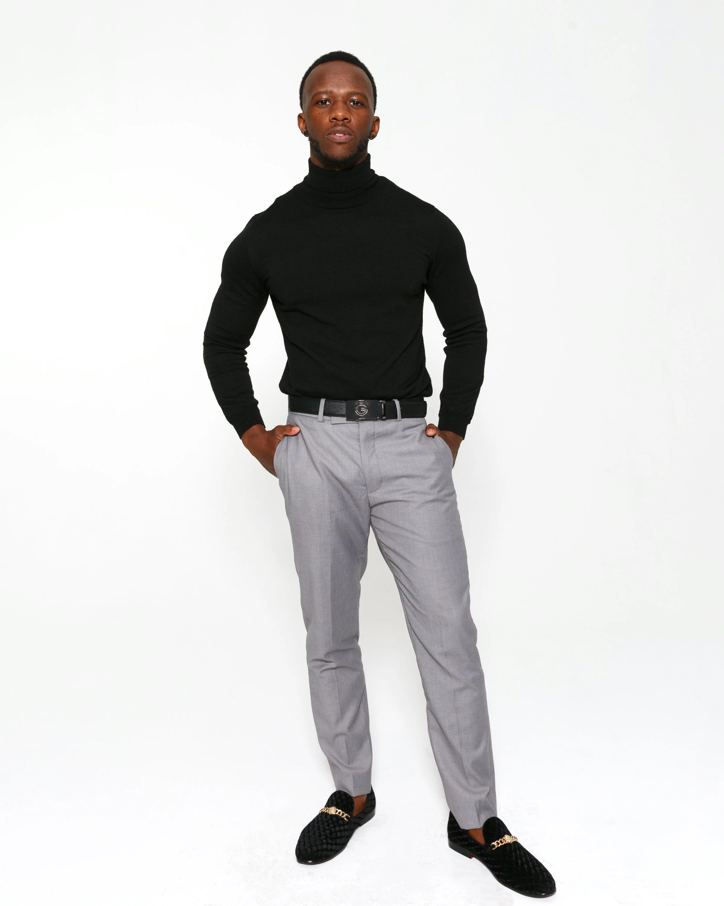 A man in a black turtleneck and gray trousers stands against a white background, with one hand in his pocket and wearing black shoes with gold chain accents.