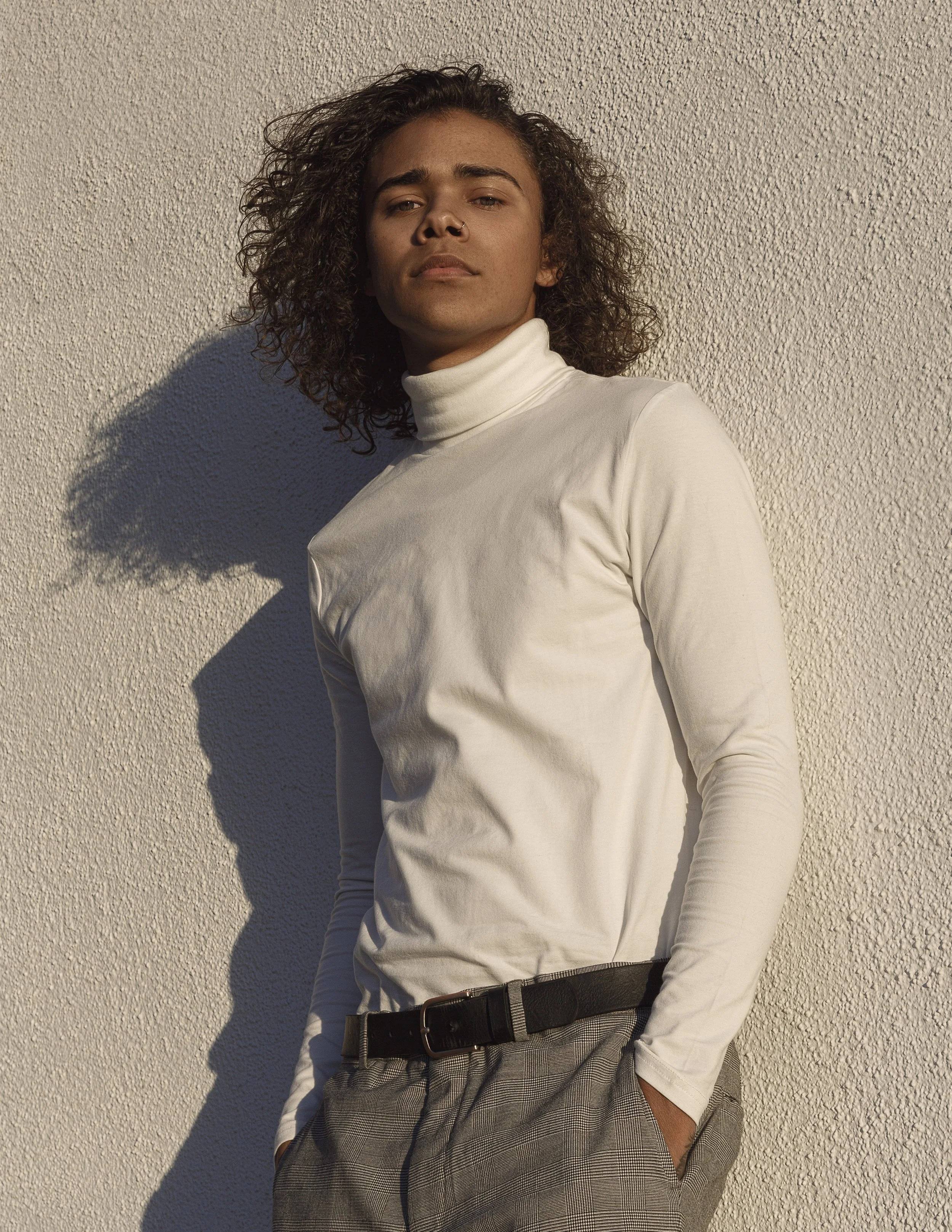 A person with curly hair and tan skin wearing a white turtleneck, black belt, and gray plaid pants, standing against a textured beige wall, looking at the camera.