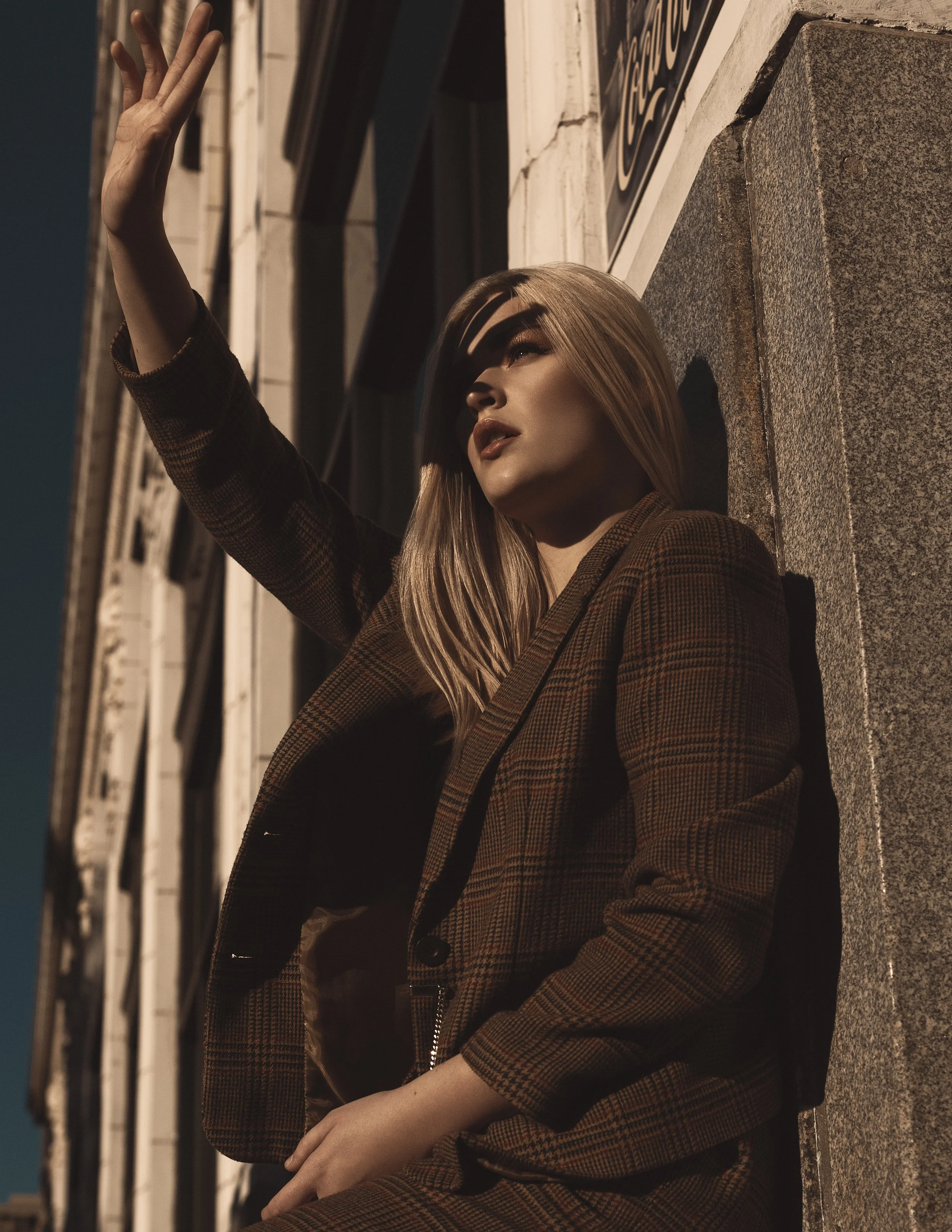 A woman with long blonde hair sitting against a building at night, wearing a brown checkered jacket, with her hand raised and shadow cast on her face.