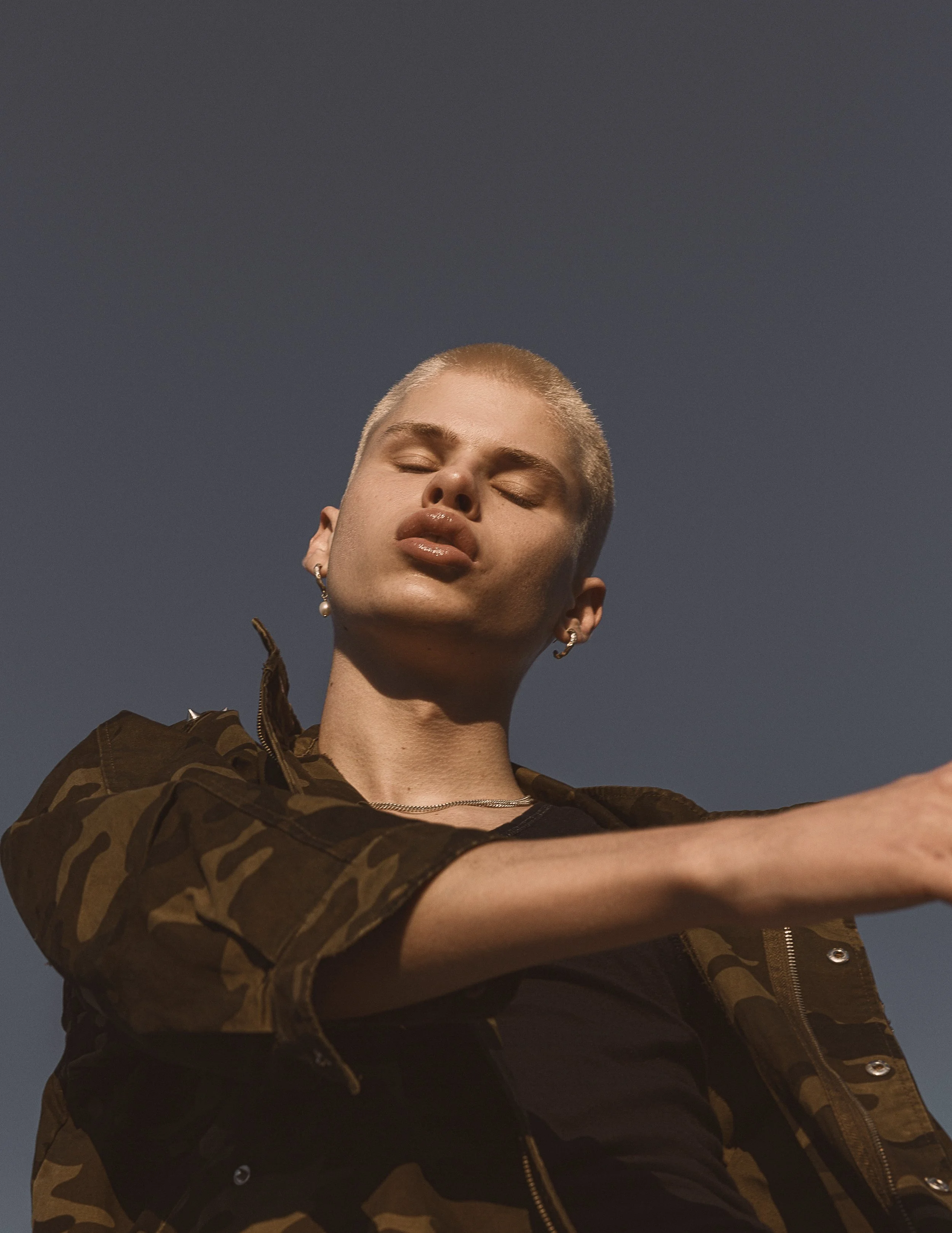 Close-up photo of a person with short blonde hair, eyes closed, wearing a camouflage jacket and earrings, against a dark sky background.