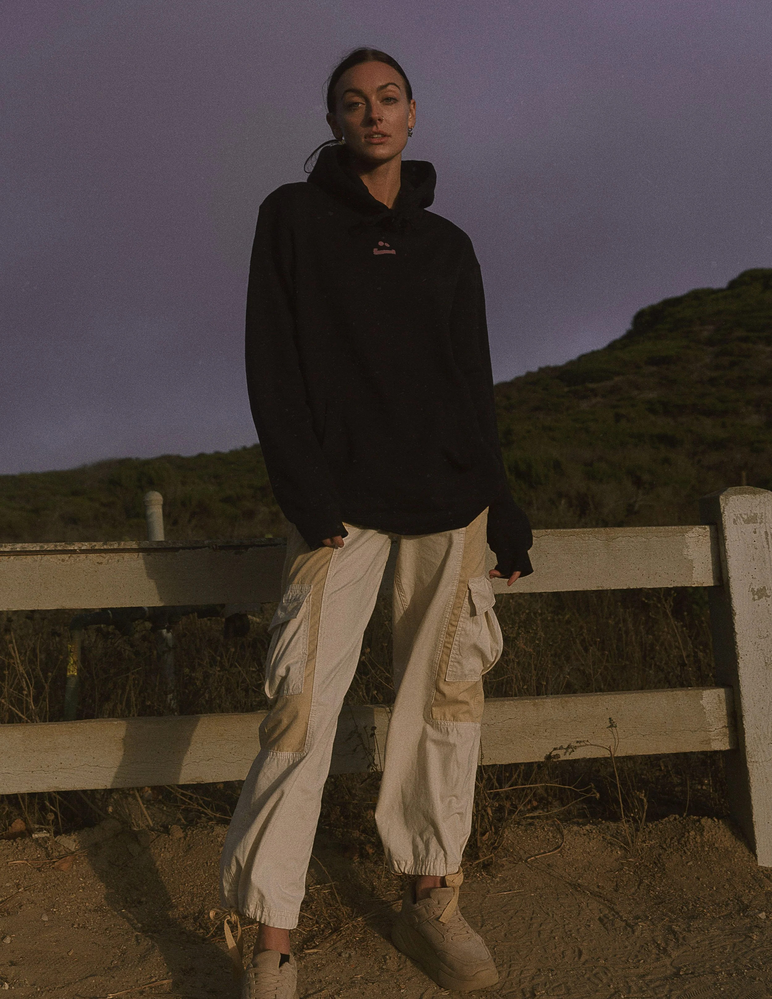A woman standing outdoors at dusk or evening, wearing a black hoodie and beige cargo pants, with a background of grassy hills and a darkening sky.