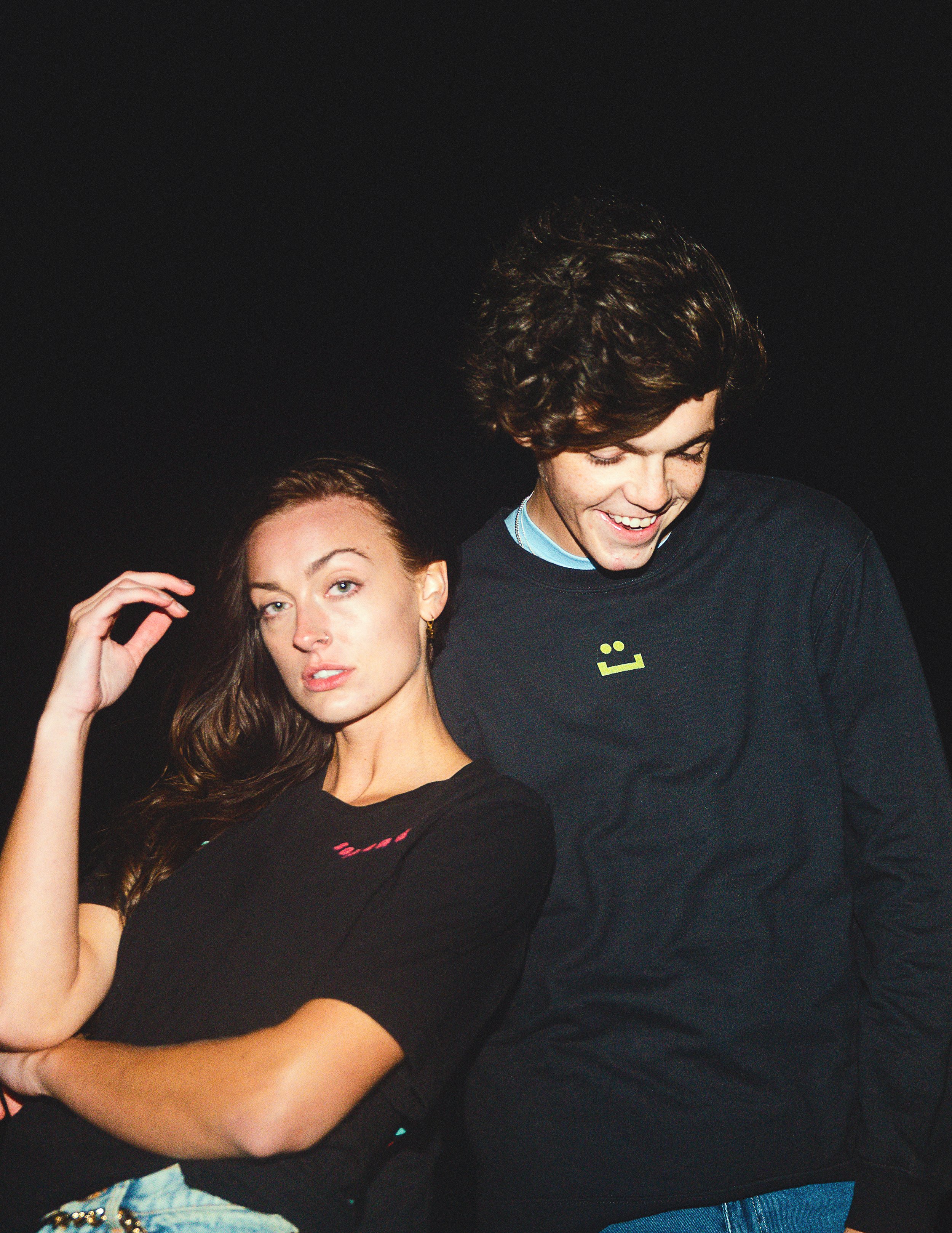 Close-up of a man smiling and looking down, and a woman with long brown hair and a nose piercing, her hand touching her hair against a black background.