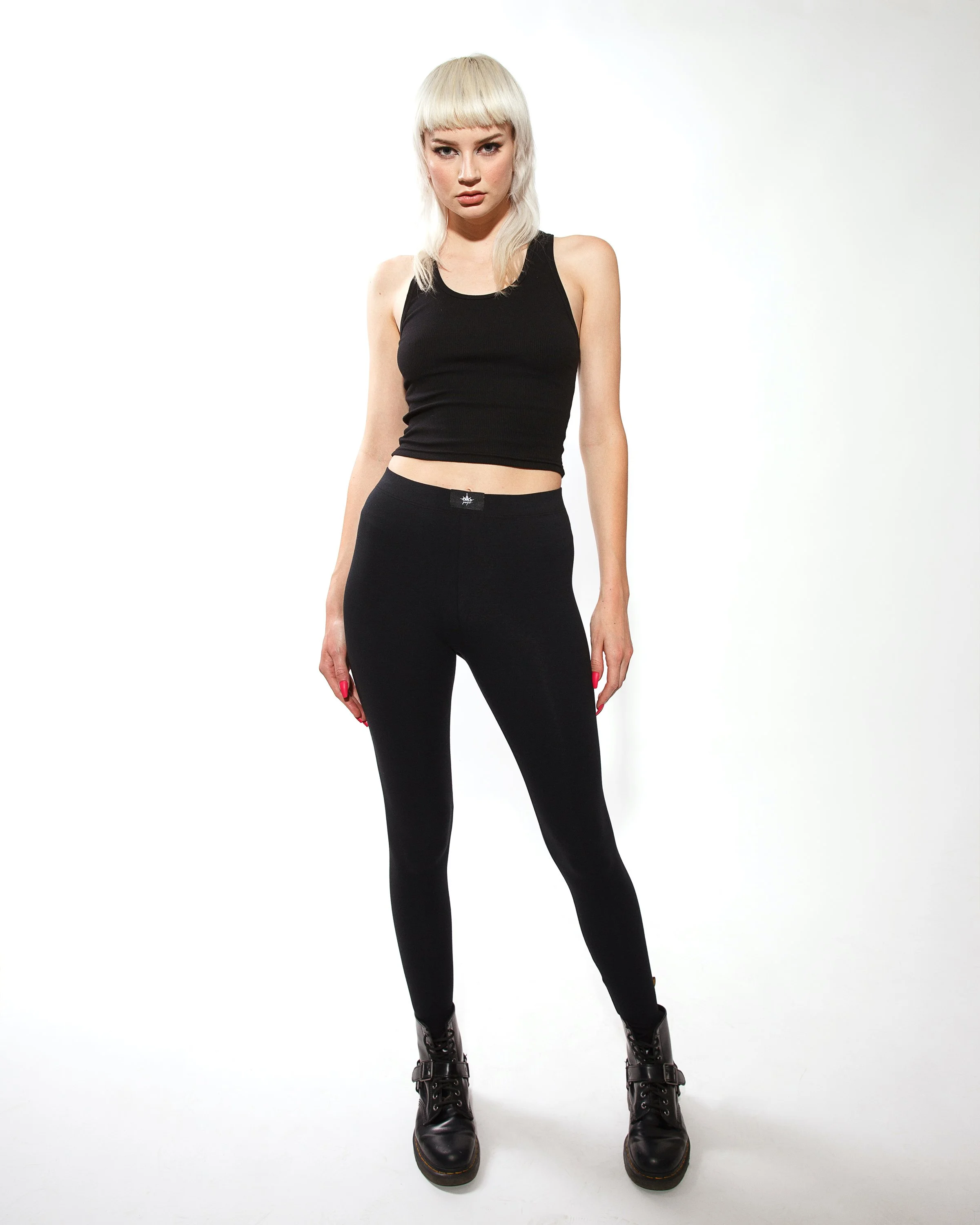 A woman with platinum blonde hair and bangs wearing a black sleeveless crop top, black high-waisted pants, and black boots standing against a plain white background.
