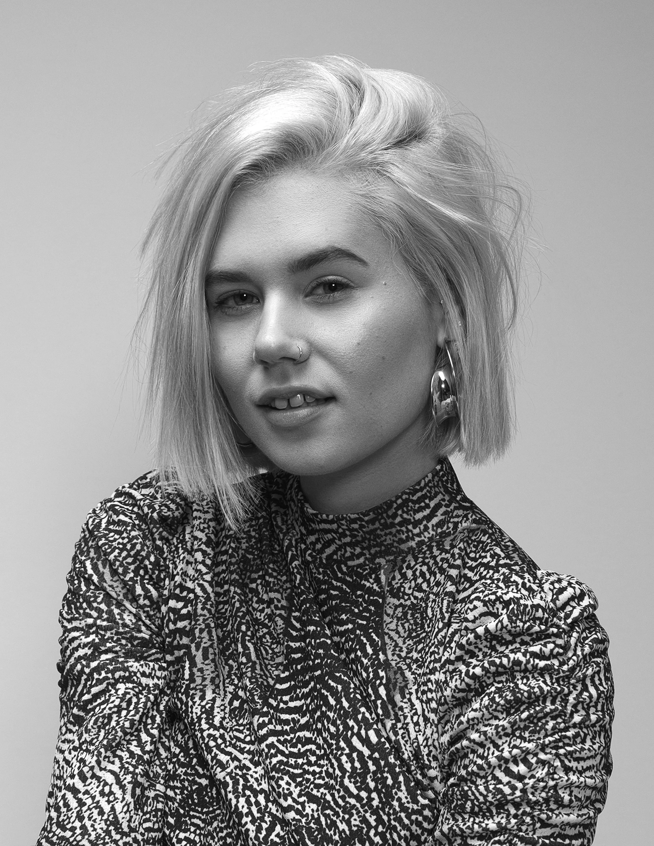 A woman with short, messy, blonde hair, wearing large earrings and a patterned shirt, smiling with her arms crossed, in black and white.
