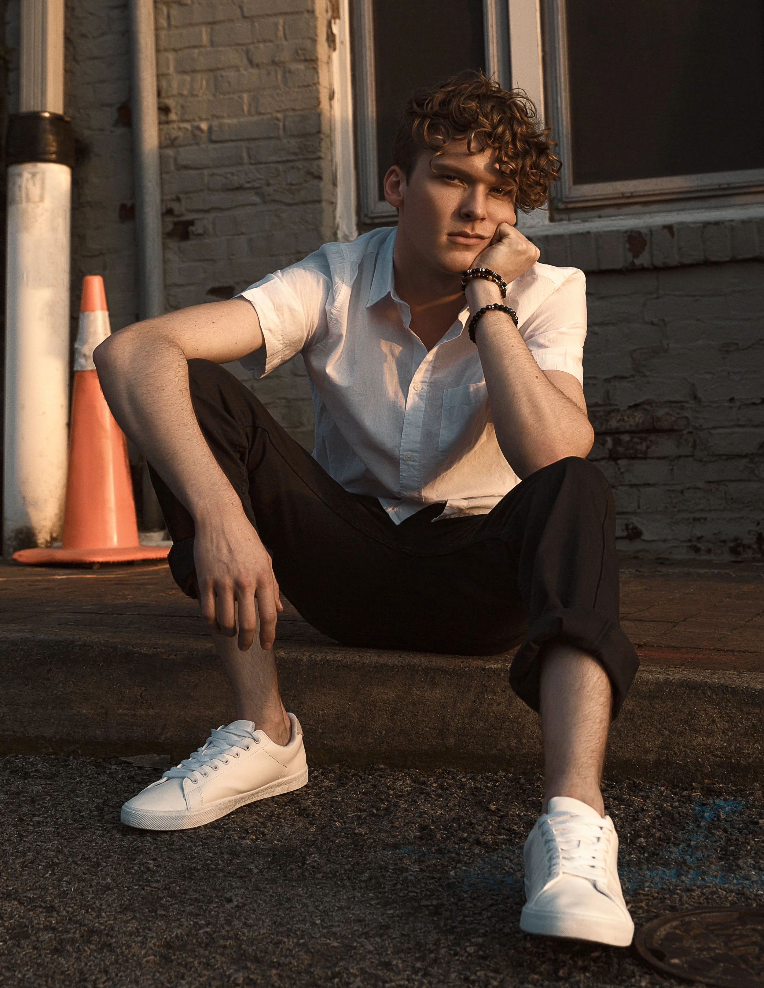 A young man with curly hair sitting on the ground outside a brick building during sunset, wearing a white shirt, black pants, and white sneakers, with a traffic cone nearby.