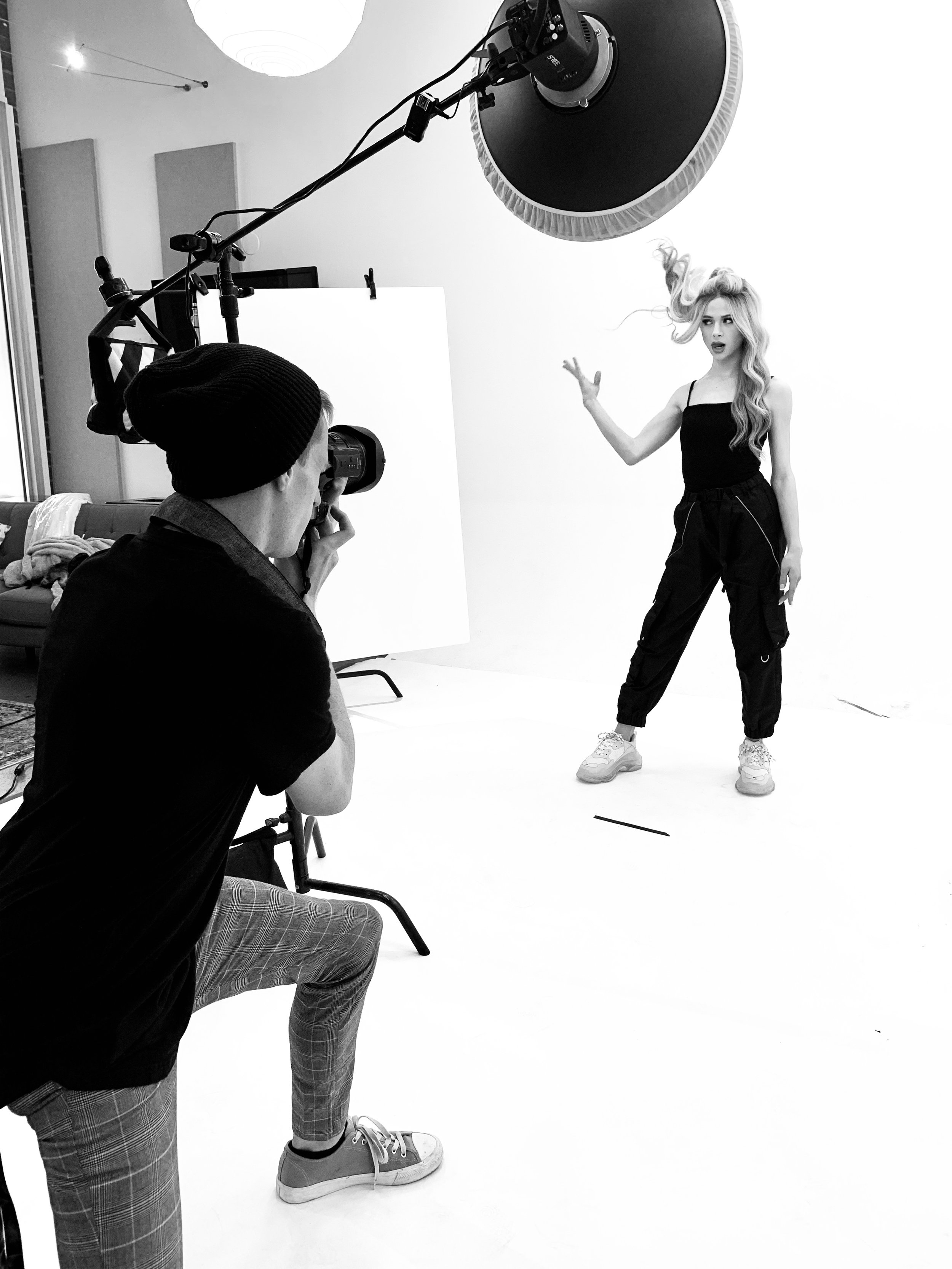 A woman with long wavy hair, wearing a black sleeveless top and black pants with white sneakers, posing in a photography studio with a white backdrop. A photographer wearing a beanie and plaid pants is taking her photo with a camera. Studio equipment includes a large softbox light overhead and a white reflector.