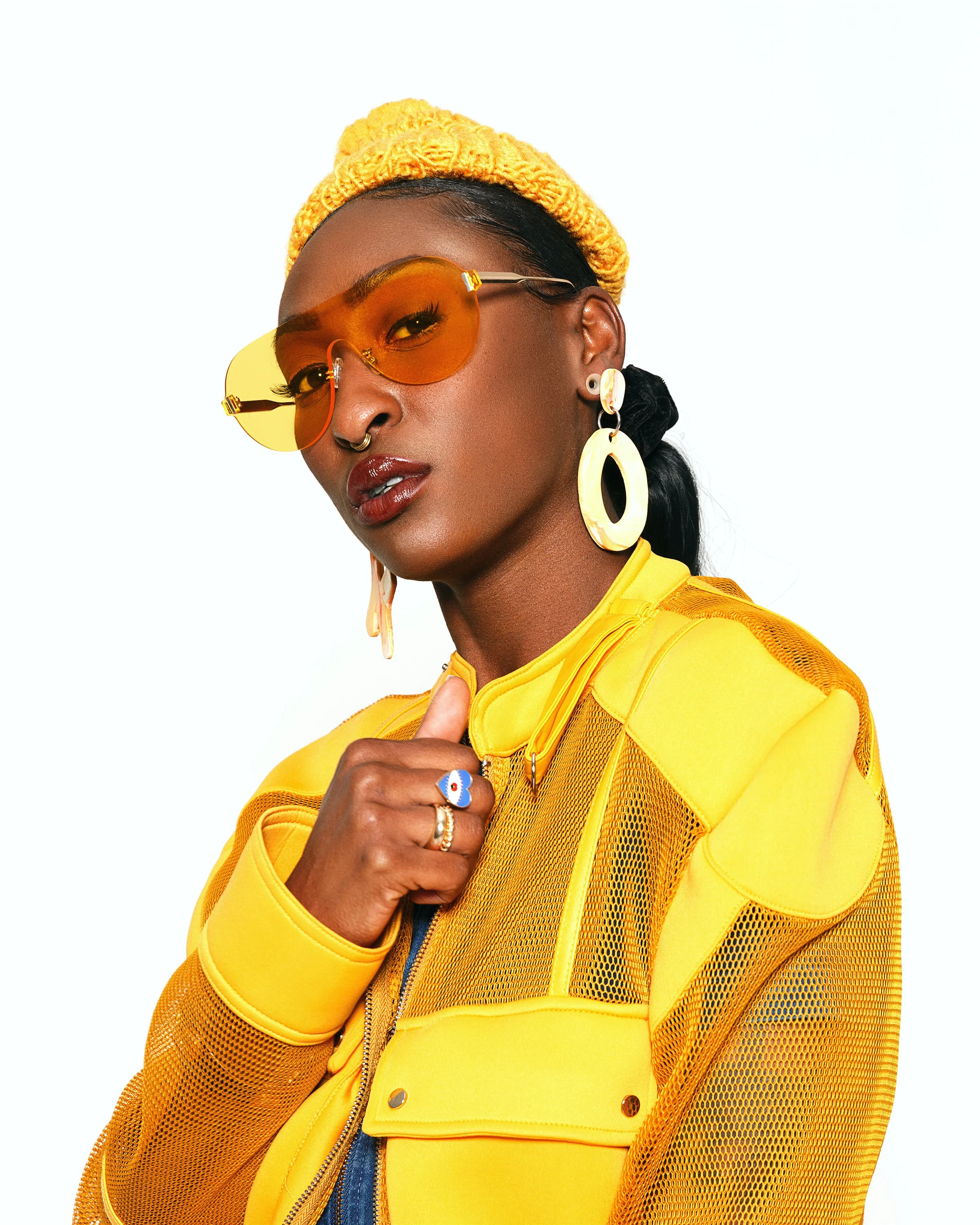 A woman with dark skin wearing yellow-tinted sunglasses, a yellow knit hat, large yellow earrings, and a yellow jacket with a zipper, against a plain white background.
