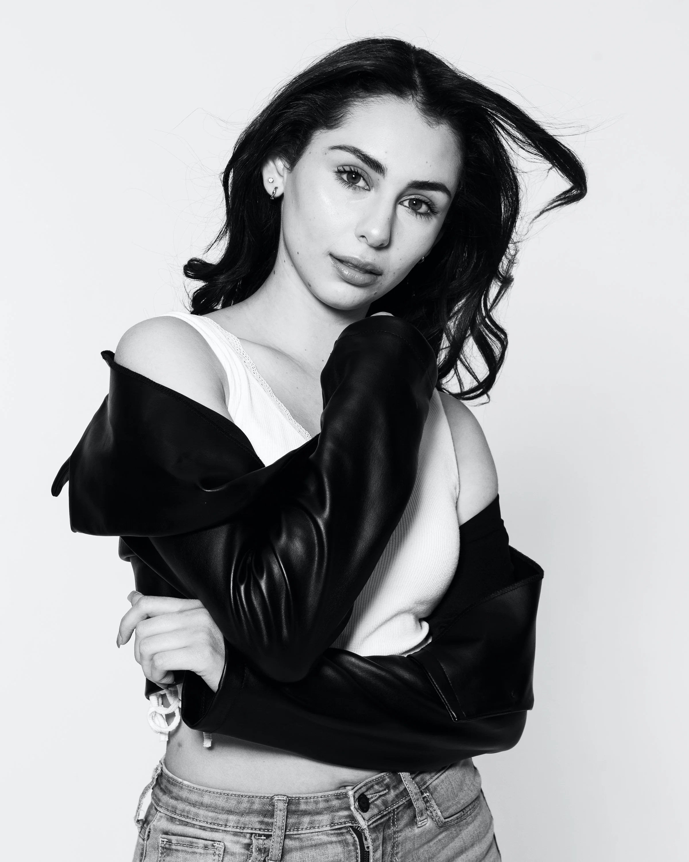 A black and white photo of a young woman with long dark hair, wearing a sleeveless top and denim shorts, with a leather jacket draped over her shoulders, standing against a plain white background.
