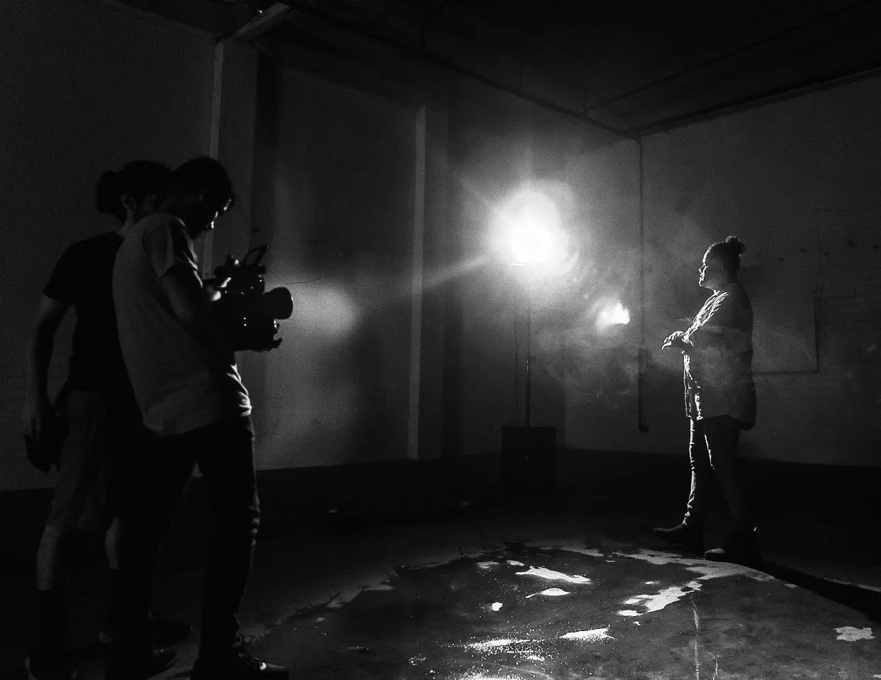 A woman speaking into a microphone while being filmed by a cameraman, in a dark room with bright lighting focused on her.