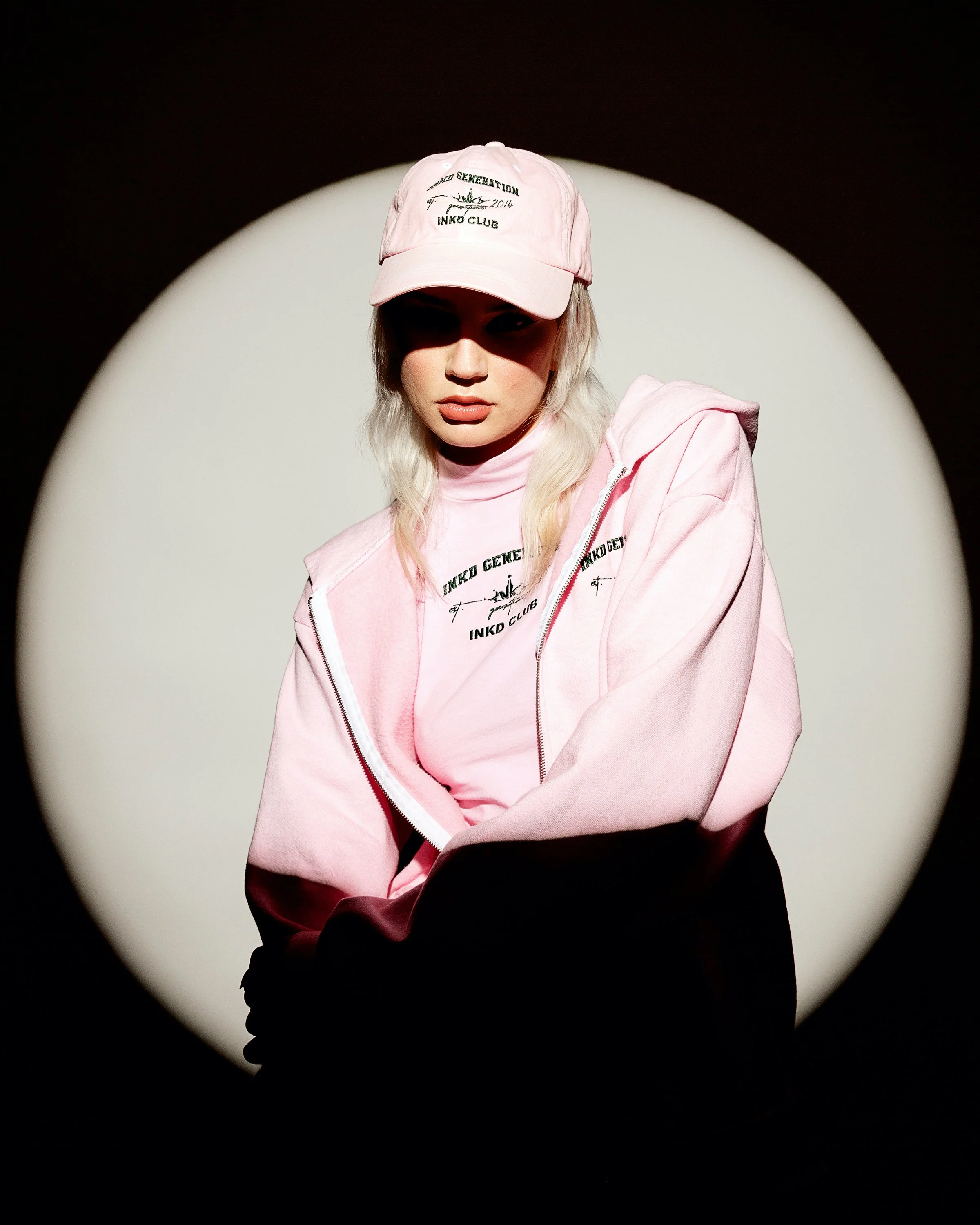 A woman with blonde hair wearing a pink baseball cap and a pink zip-up hoodie, standing in a spotlight against a dark background.